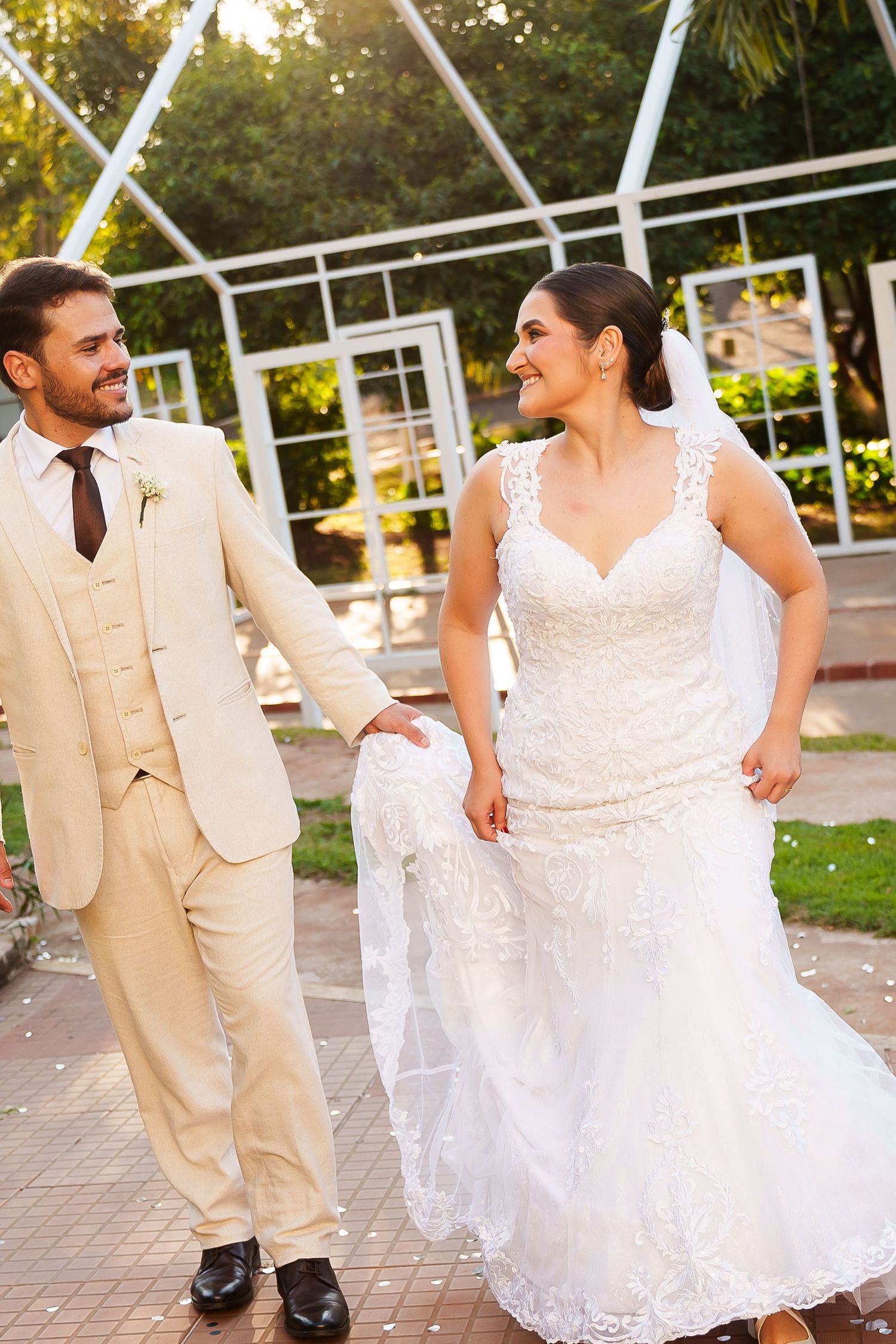 A smiling bride and groom walk hand in hand outdoors, she in a lace gown and he in a beige suit, at a garden wedding.