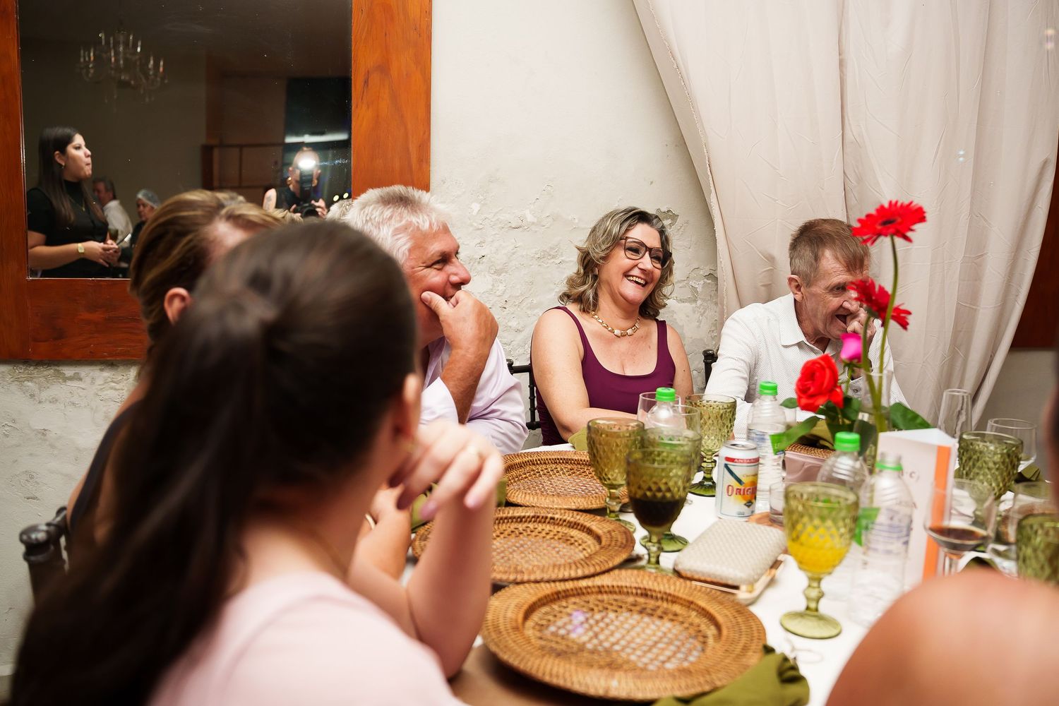 Group of adults laughing and socializing at a festive dinner table with green glassware and floral centerpiece.