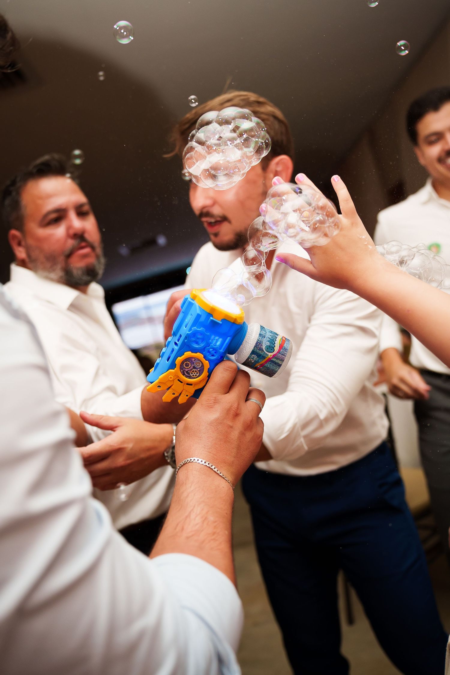 Men in white shirts celebrate with a bubble gun toy at a party, covered in soap bubbles indoors.