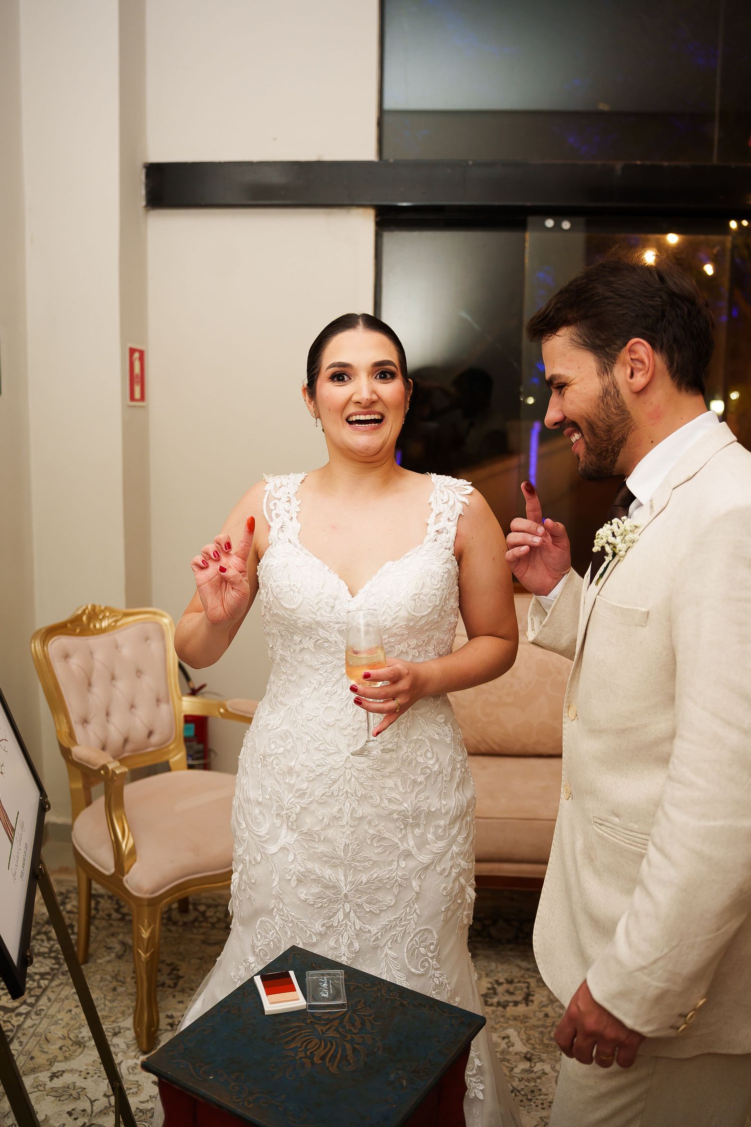 Bride in lace gown laughing with groom in white suit at elegant wedding reception holding champagne.