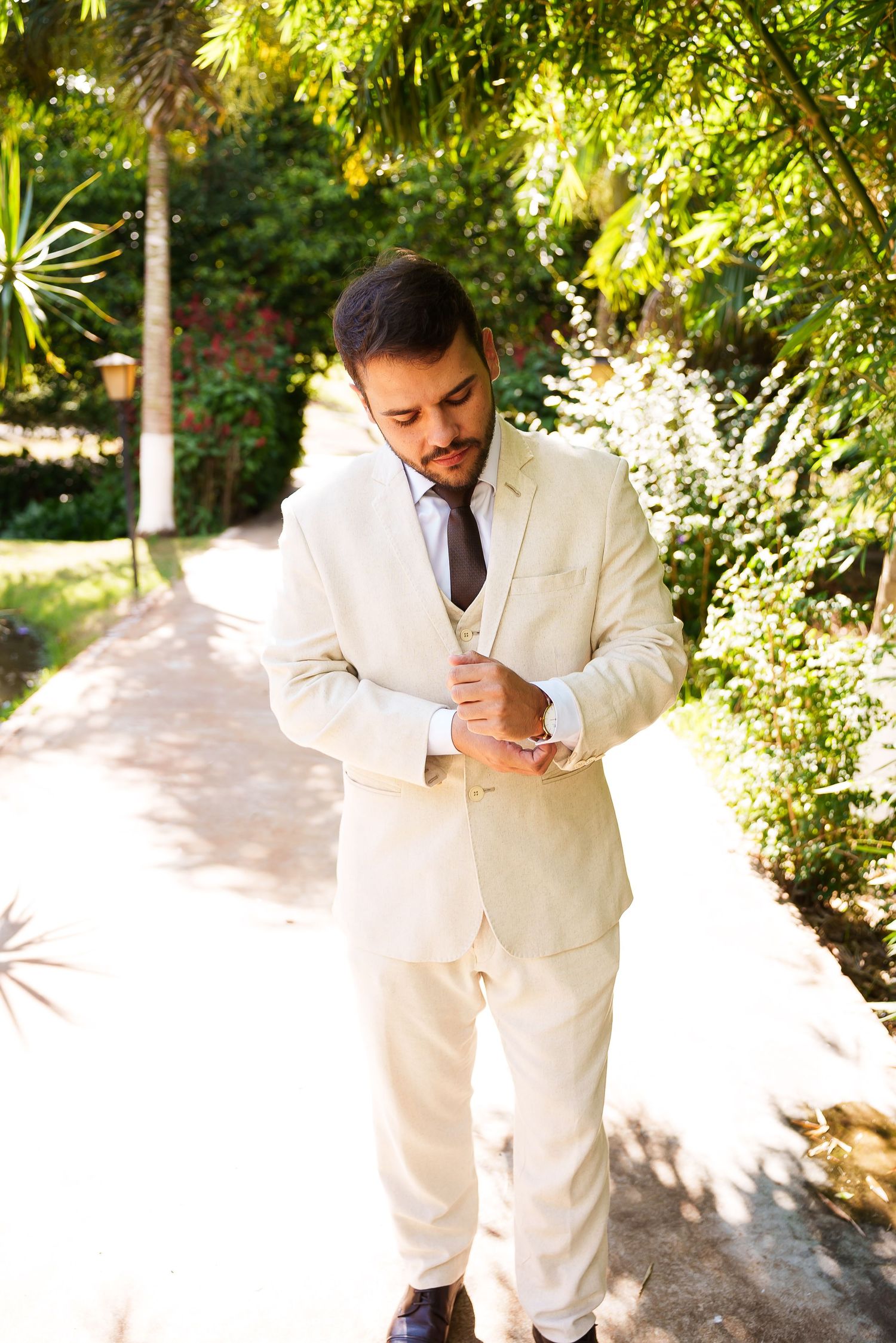 A well-dressed groom in a cream three-piece suit adjusts his cufflinks on a sunlit garden path surrounded by lush greenery.