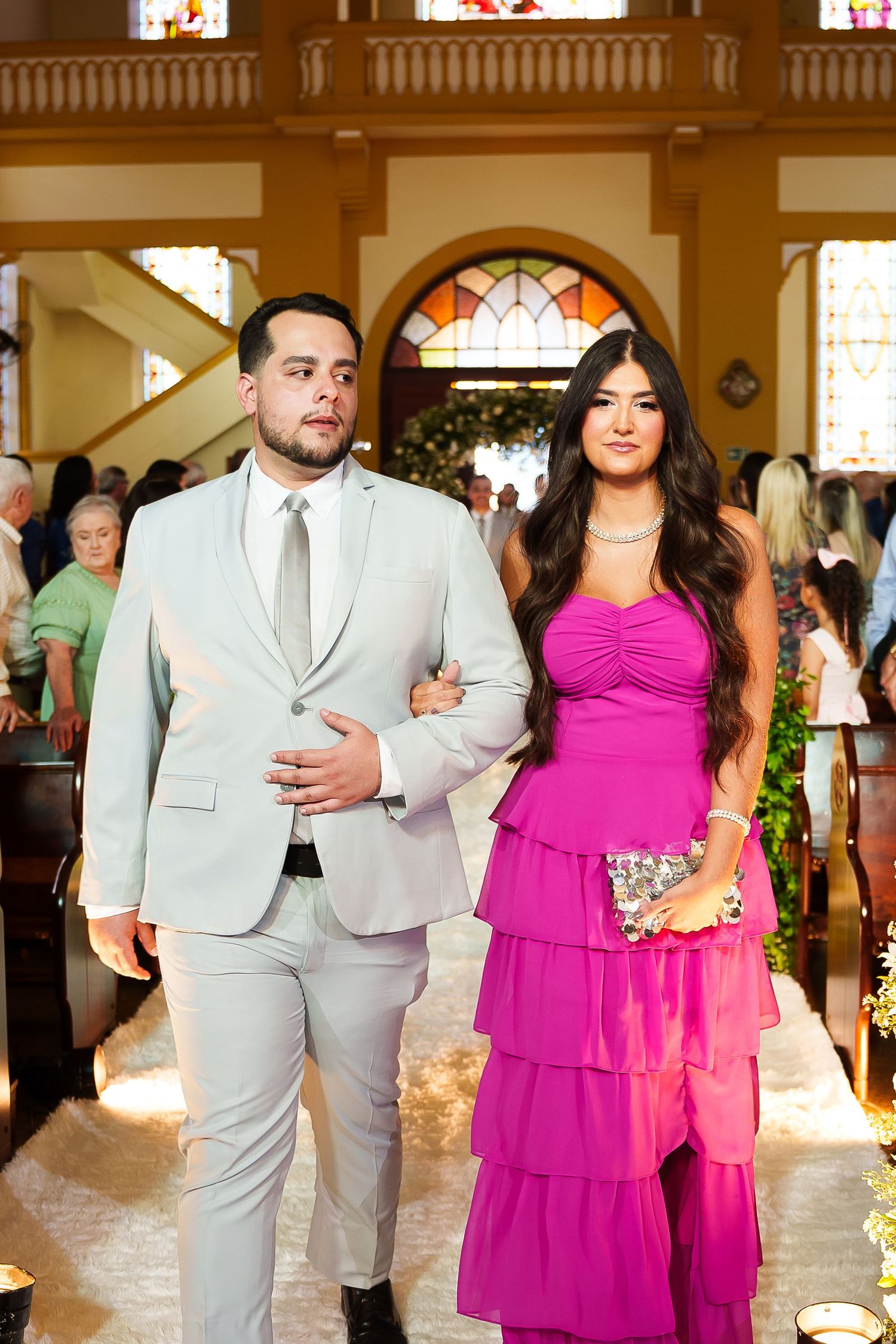 Elegant couple walking down church aisle, man in light gray suit, woman in vibrant pink tiered gown.