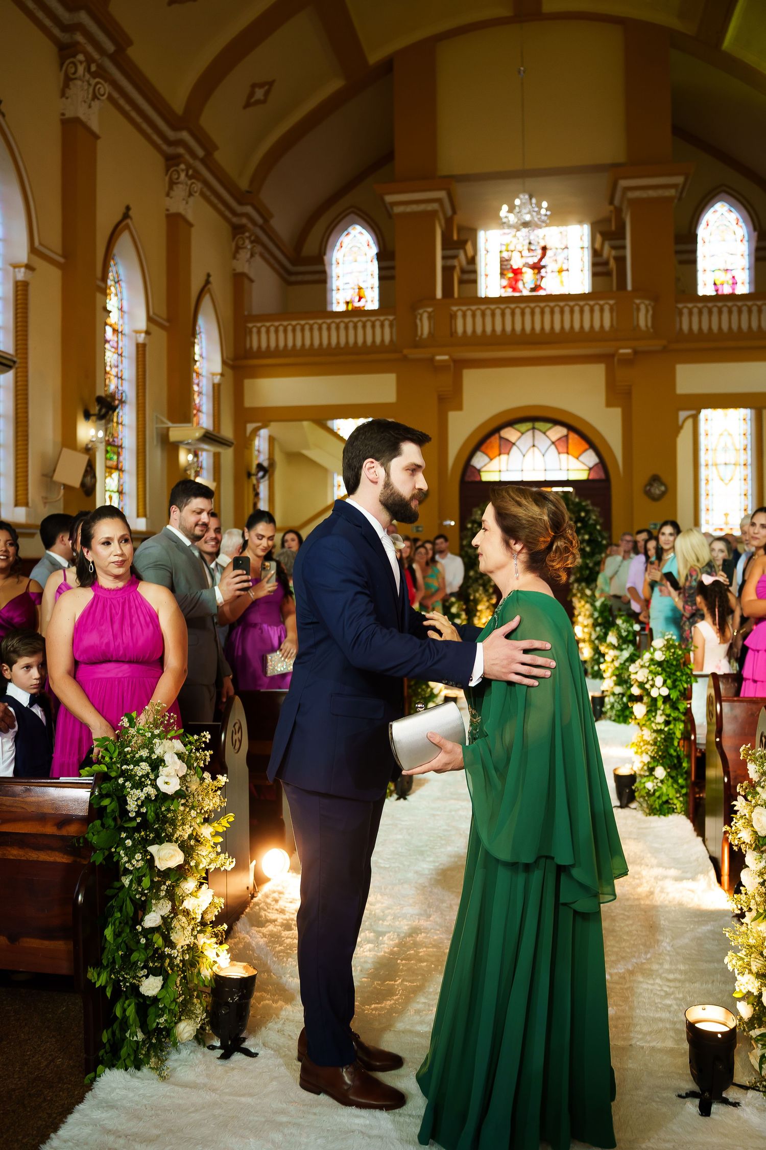 Elegant church wedding ceremony with groom in navy suit greeting bride in green gown along floral aisle.
