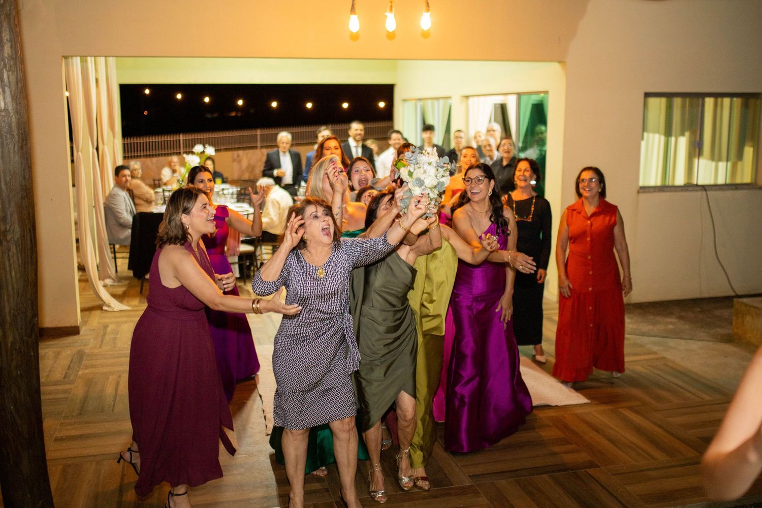 Excited wedding guests in colorful dresses reaching up to catch the bridal bouquet toss at a festive indoor reception.