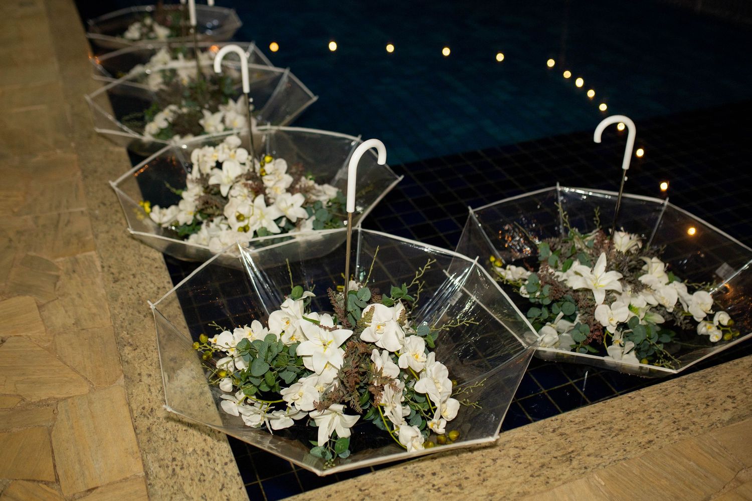 Clear umbrellas filled with white floral arrangements displayed poolside at an elegant evening event.