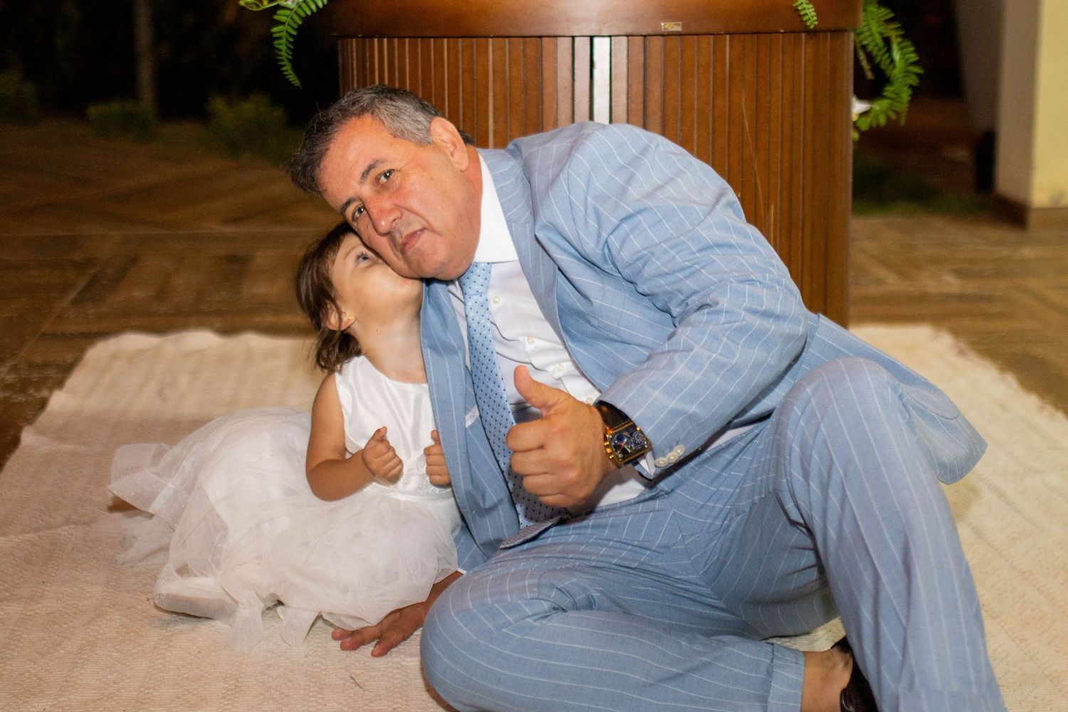 Man in blue suit and young girl in white dress share a joyful hug, both giving thumbs up at a formal event.