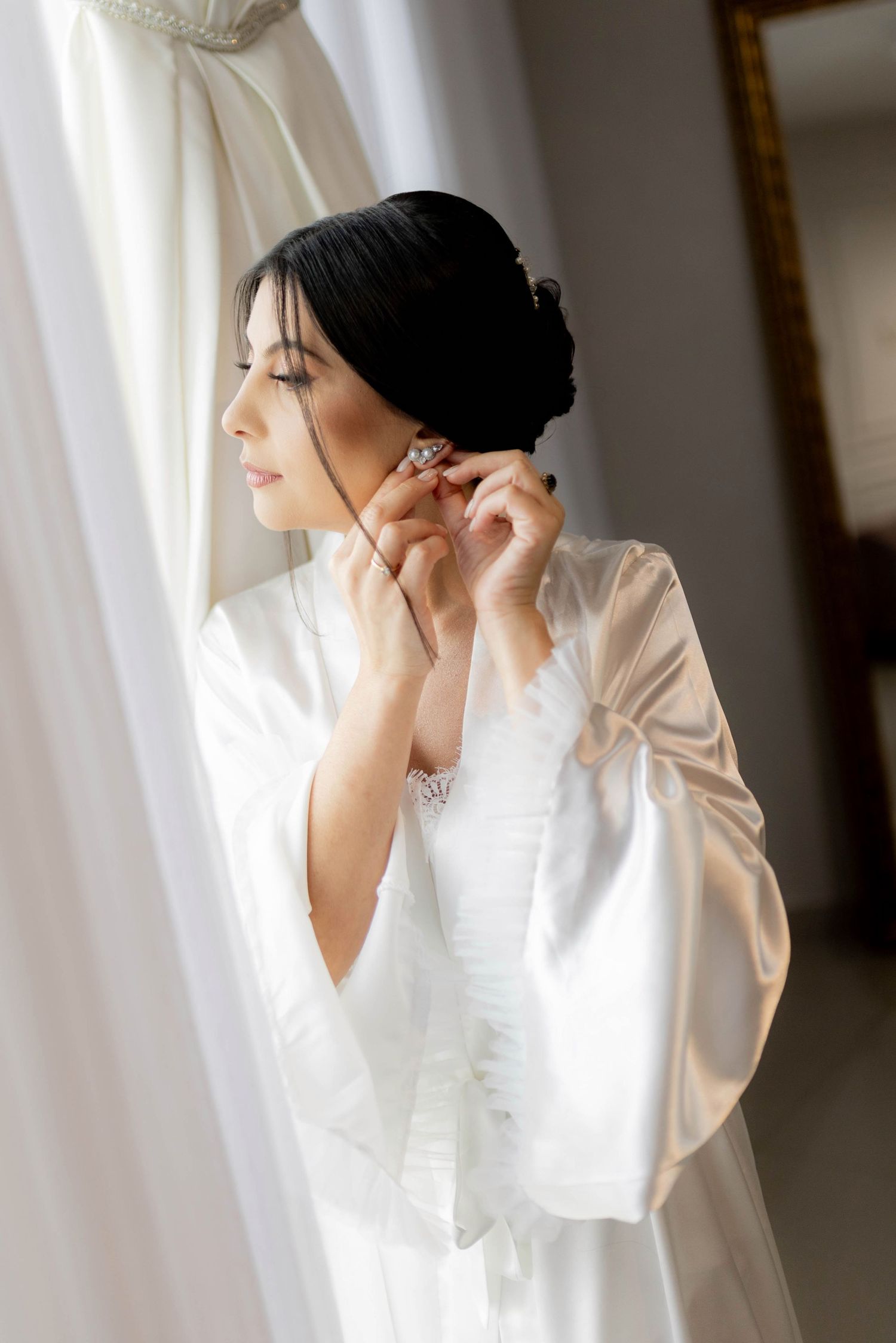 Elegant bride in white satin robe putting on crystal earrings near a window on her wedding day.