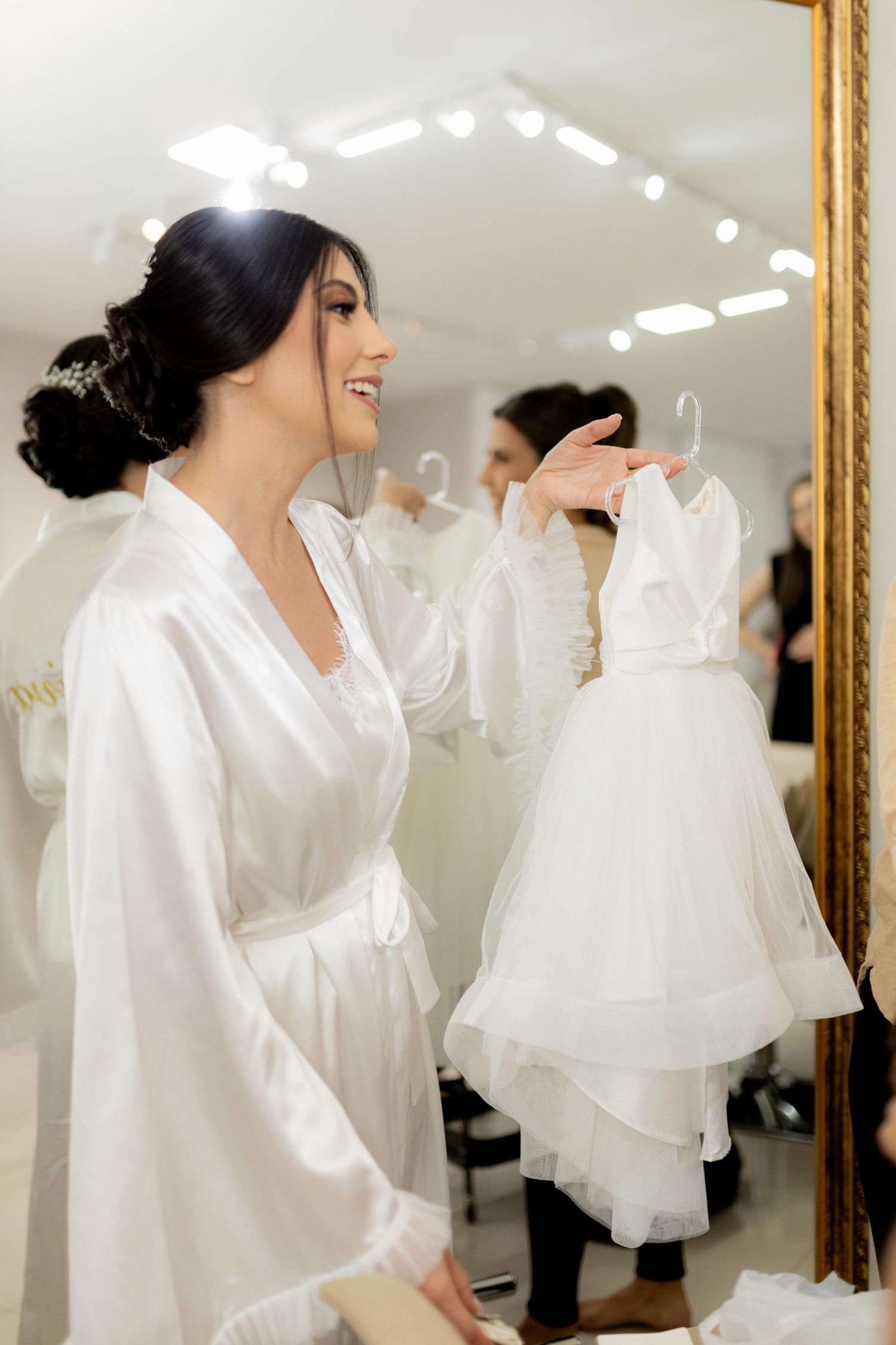 Smiling bride in white robe holds a flower girl dress up to a gold-framed mirror in a bridal boutique.