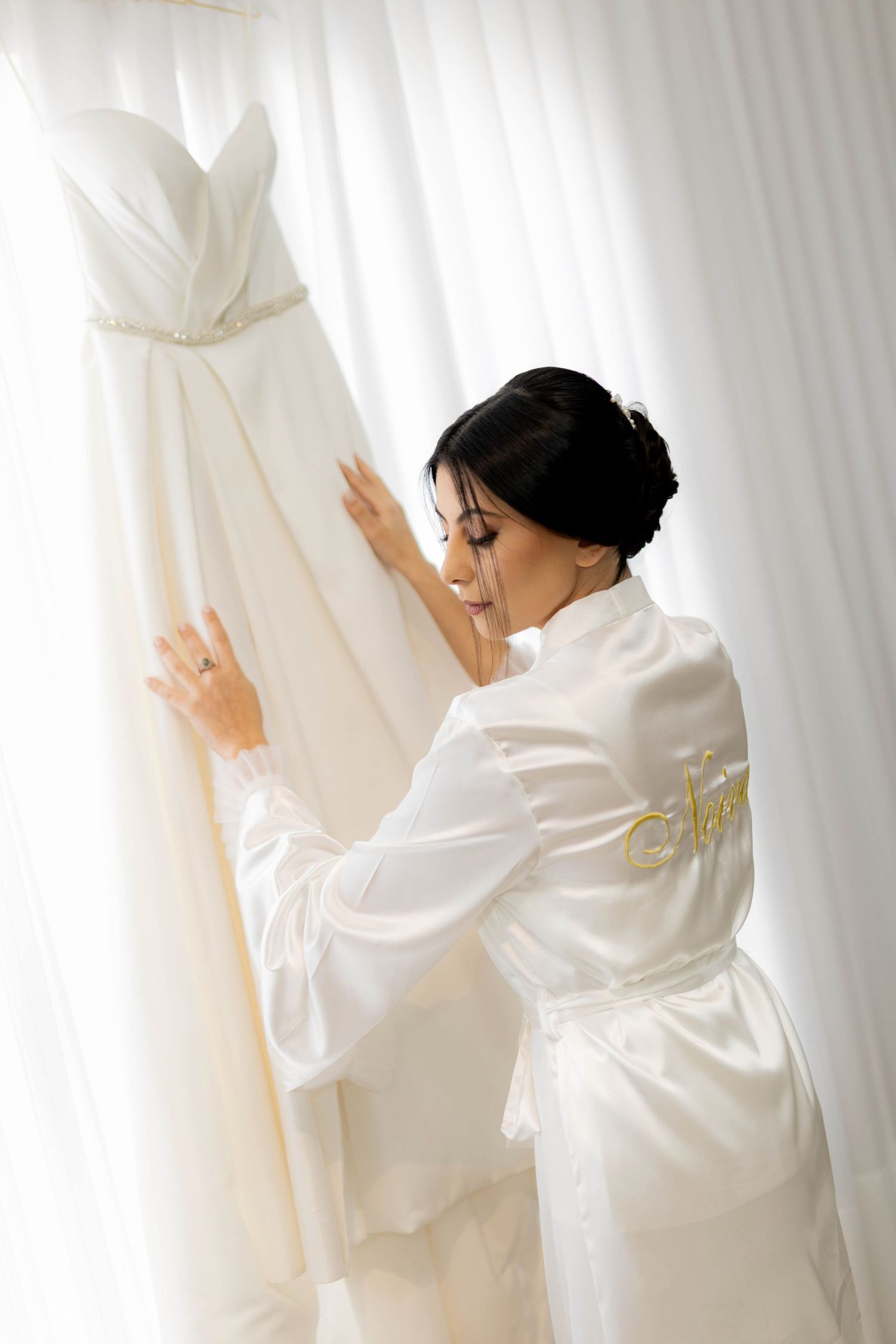 Bride in white satin robe admiring her wedding dress hanging by sheer curtains.