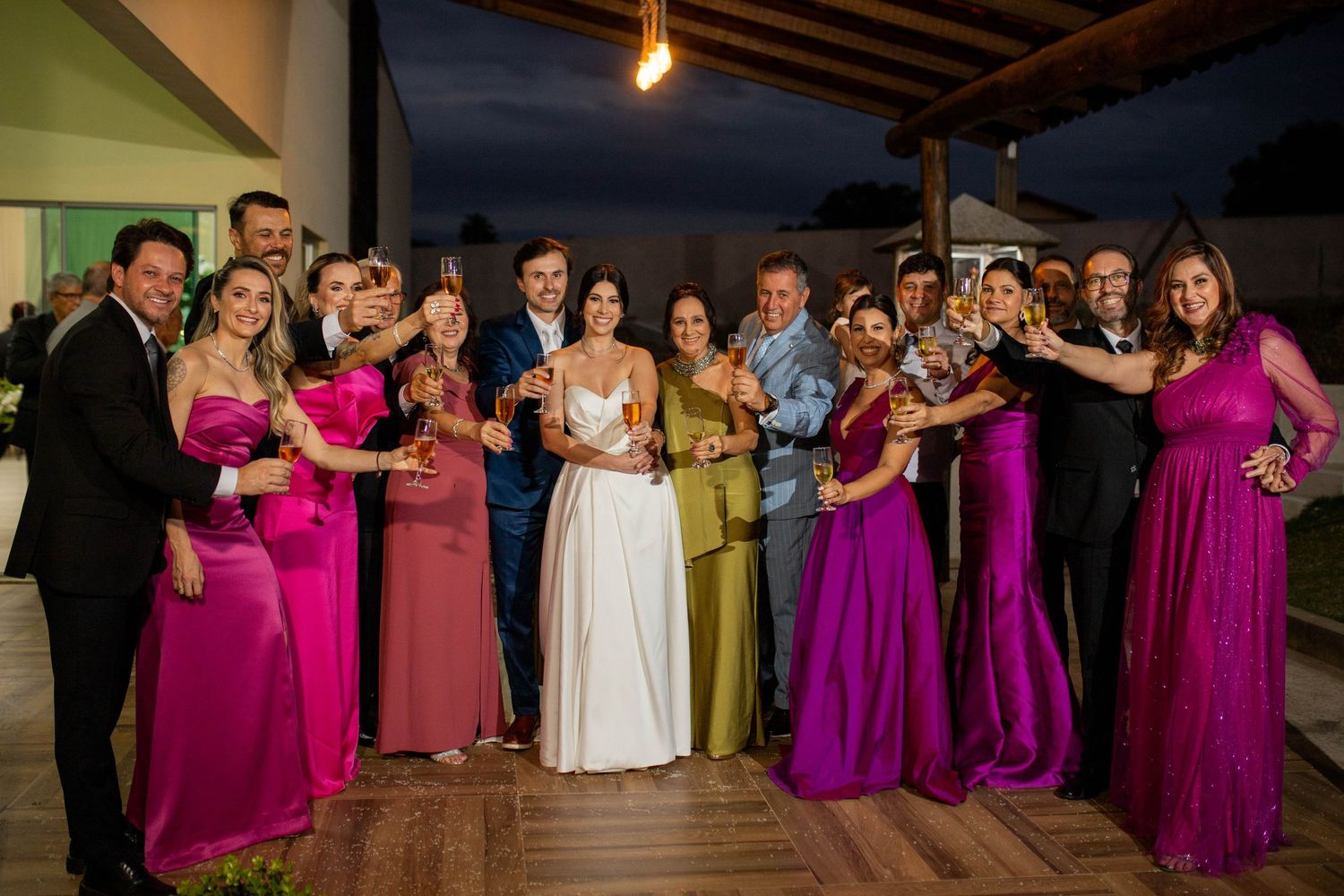 Wedding party group photo with bride in white gown, groom, bridesmaids in pink and purple dresses, toasting champagne outdoors.