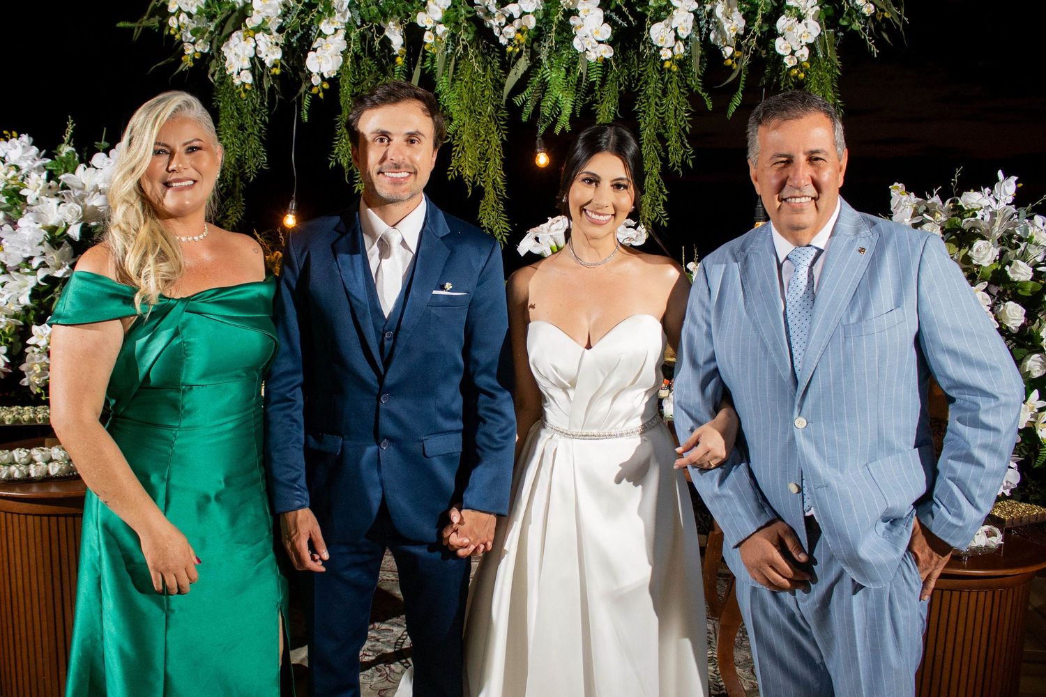 Wedding party of four posing together, bride in white gown, groom in navy suit, guests in green dress and blue suit.