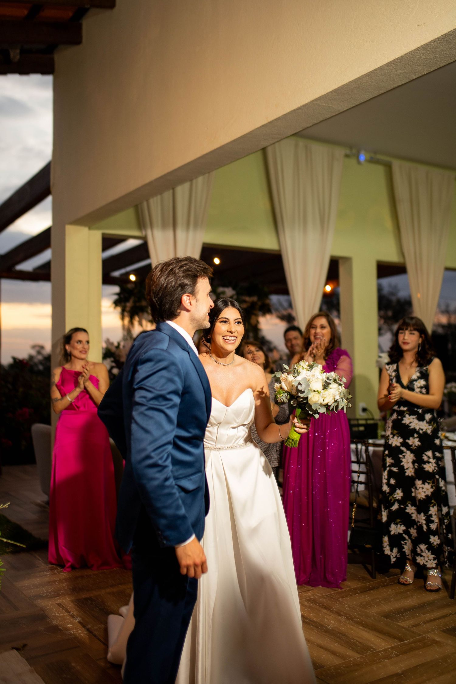 Bride in white gown and groom in blue suit share first dance at outdoor wedding reception with guests applauding.