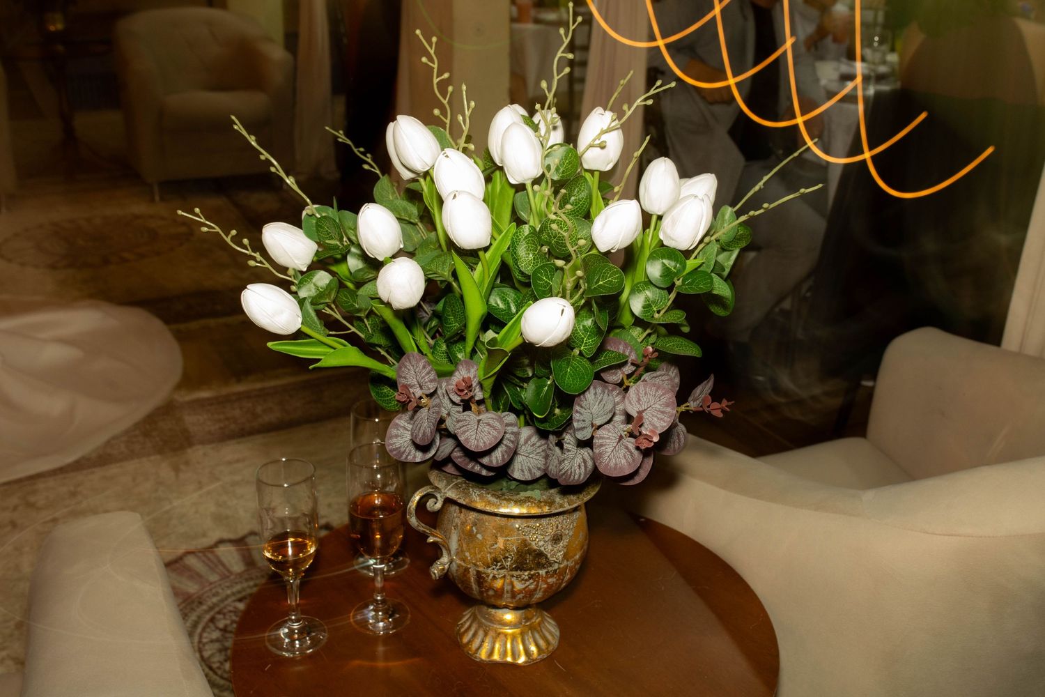 White tulips and purple flowers in a gold vase on a wooden table with wine glasses nearby.