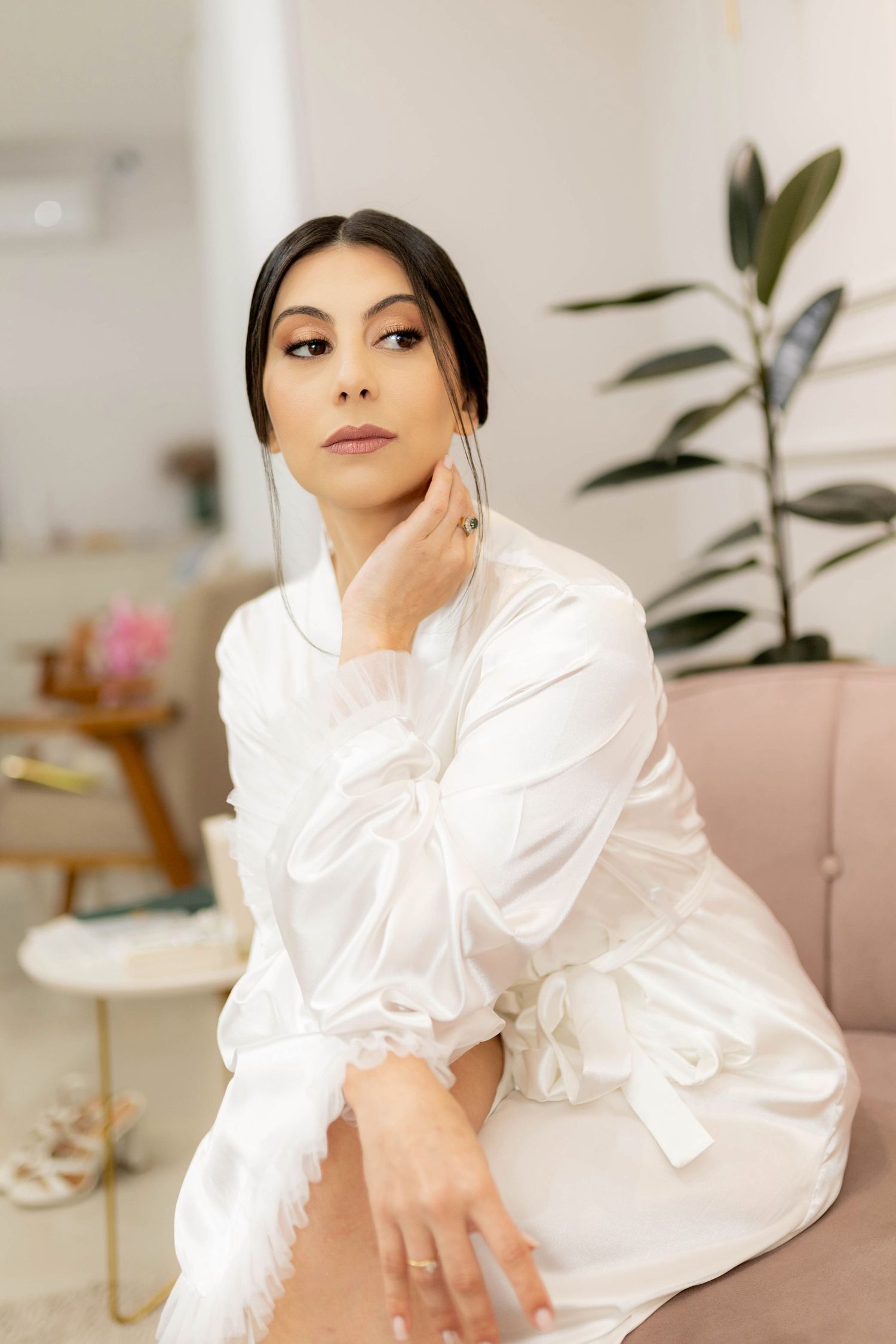A woman in a white satin robe sits elegantly, touching her face, with a plant and soft decor in the background.