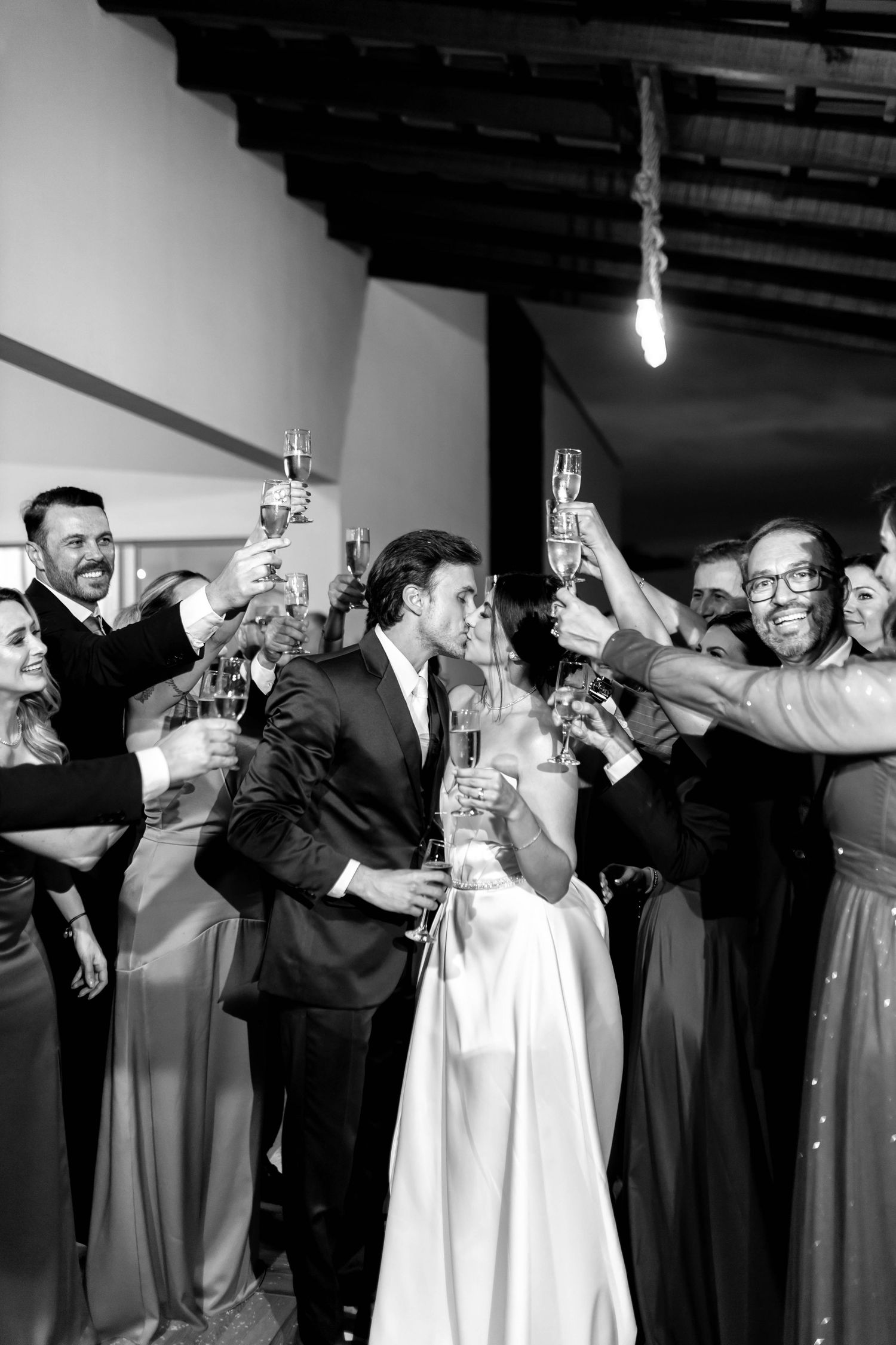 Black and white photo of bride and groom kissing during champagne toast surrounded by wedding guests.