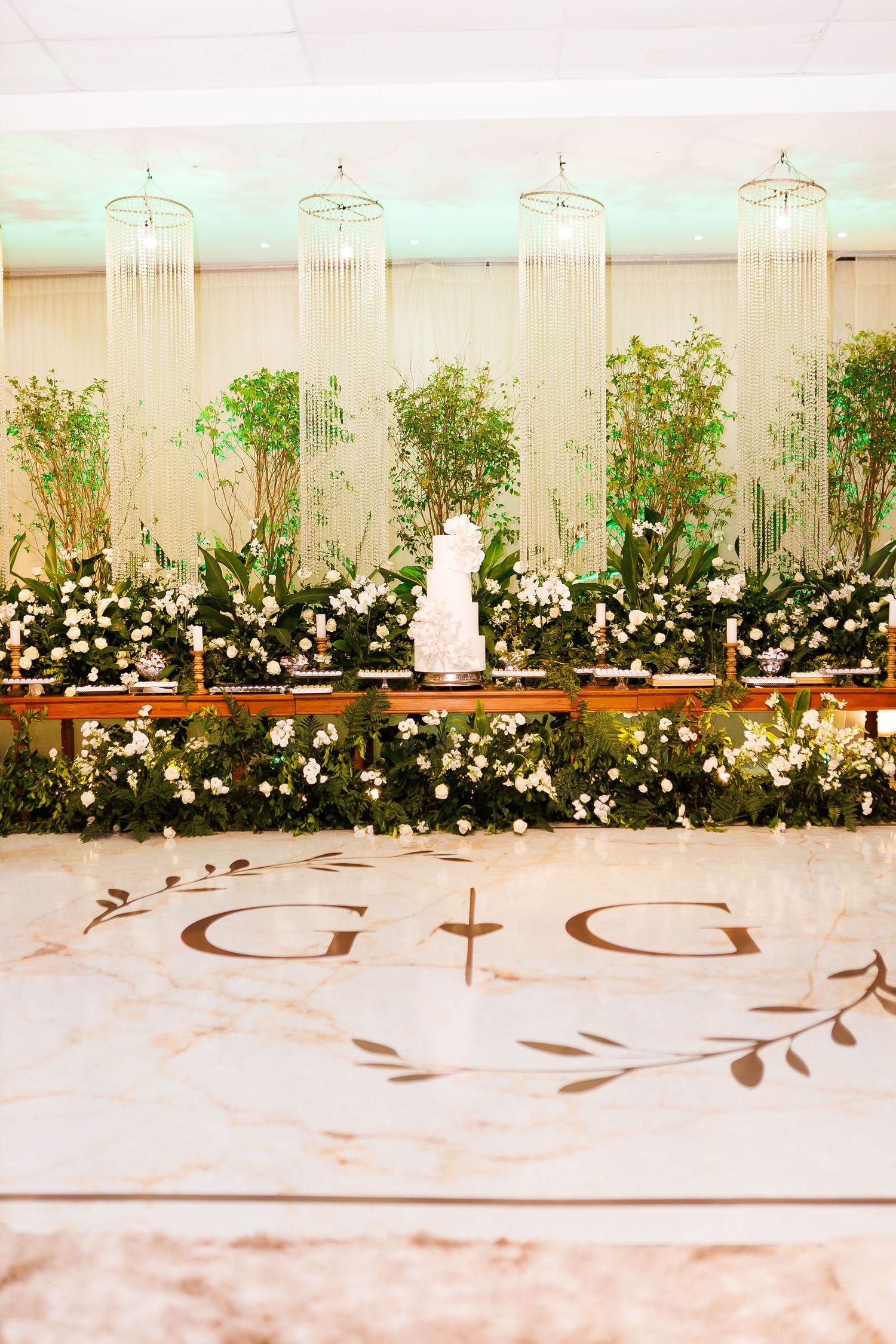 Elegant wedding reception with monogram dance floor, white tiered cake, lush greenery backdrop, candles, and crystal chandeliers.