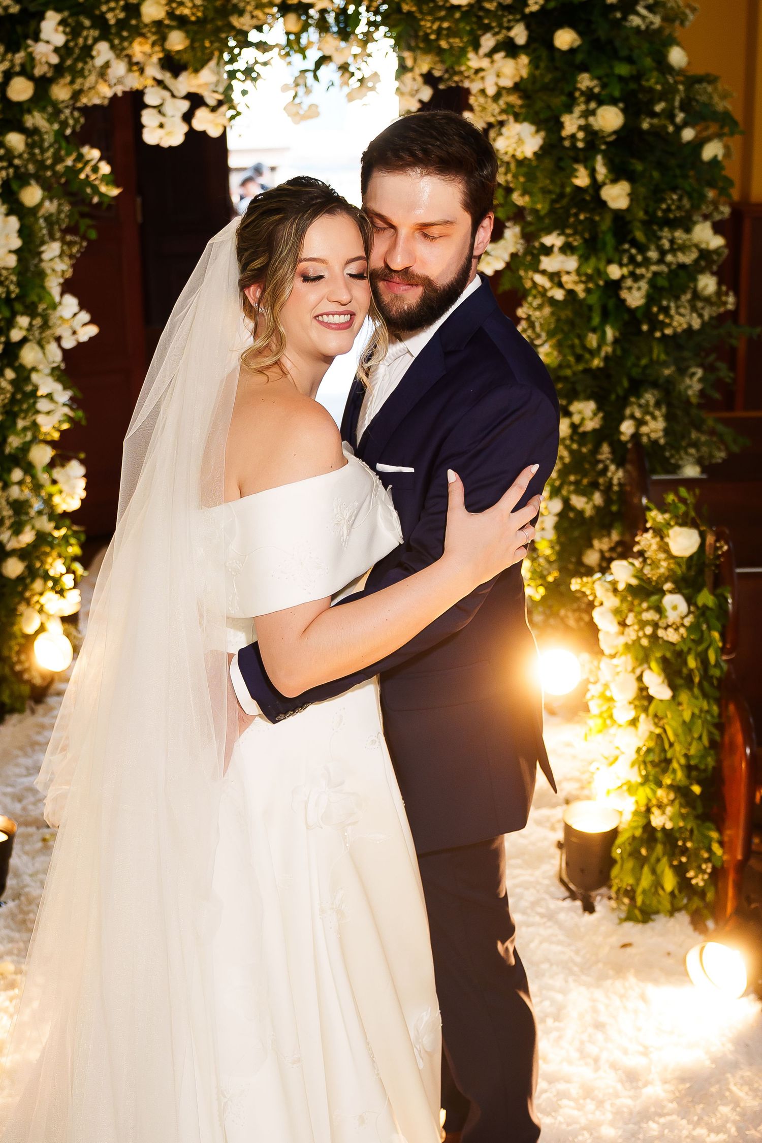 Bride and groom embracing at wedding altar adorned with white flowers and warm lights, bride wearing off-shoulder gown and veil.