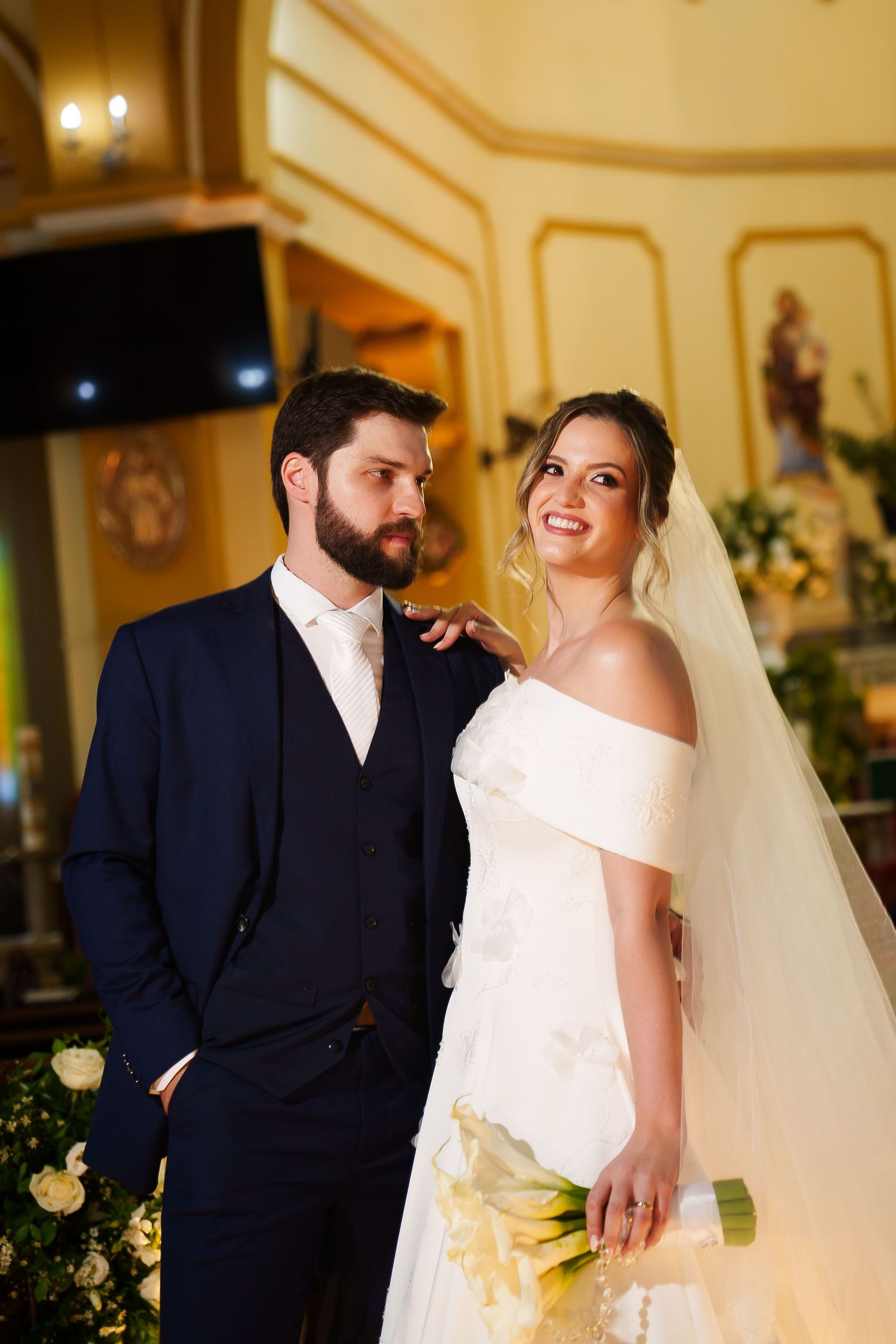 Bride in white off-shoulder gown holding bouquet stands with bearded groom in navy suit inside ornate church.