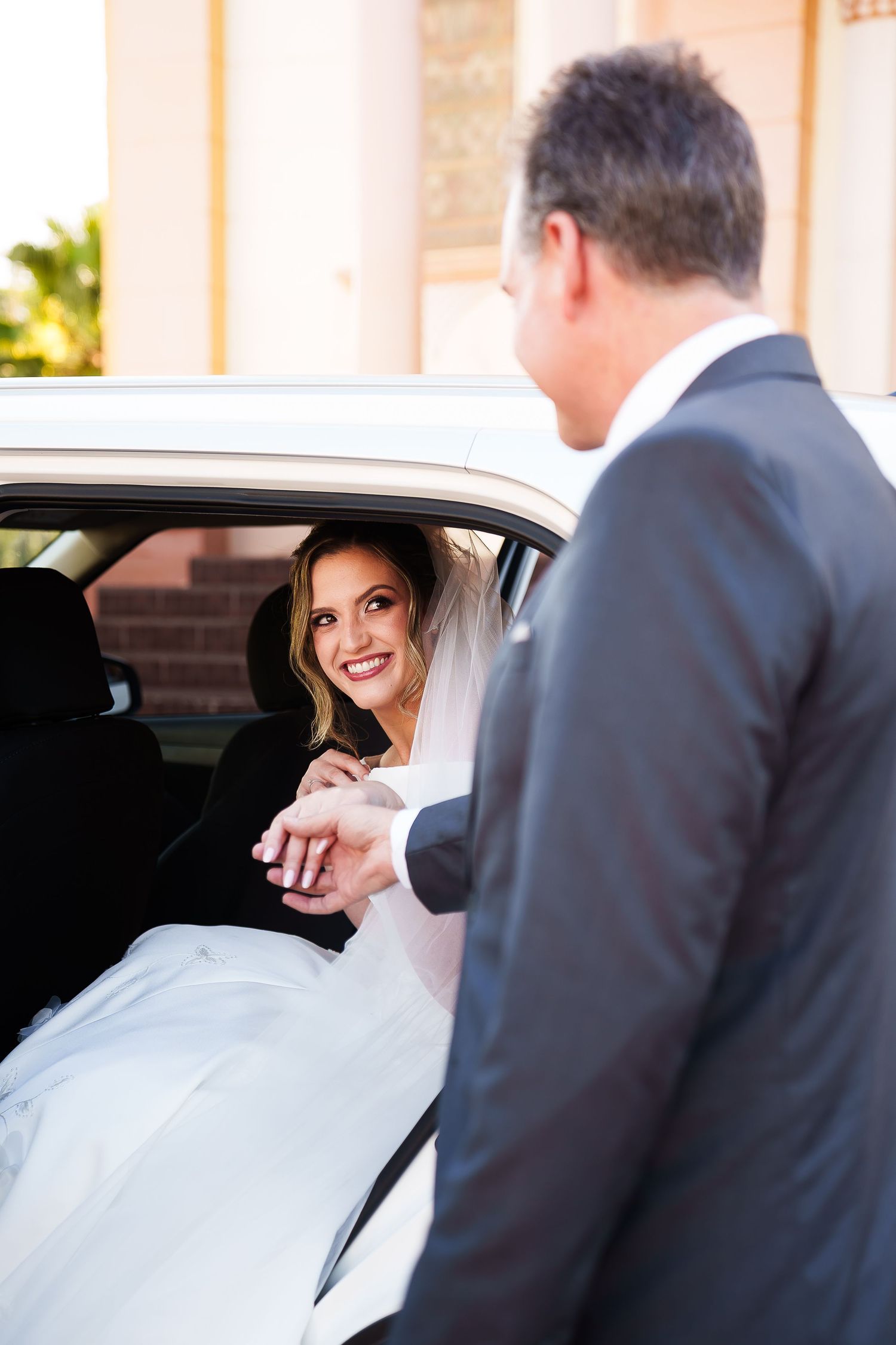 Smiling bride in white dress and veil exits car, holding hands with groom in gray suit outside church.