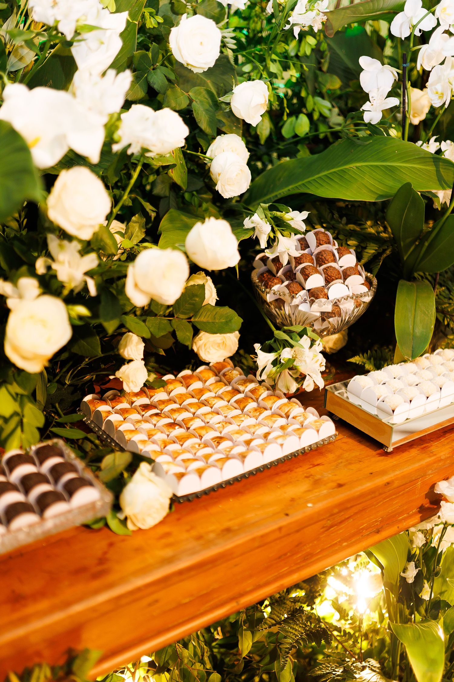 Elegant wedding candy bar with assorted chocolates and sweets on wooden table adorned with white roses and tropical greenery.