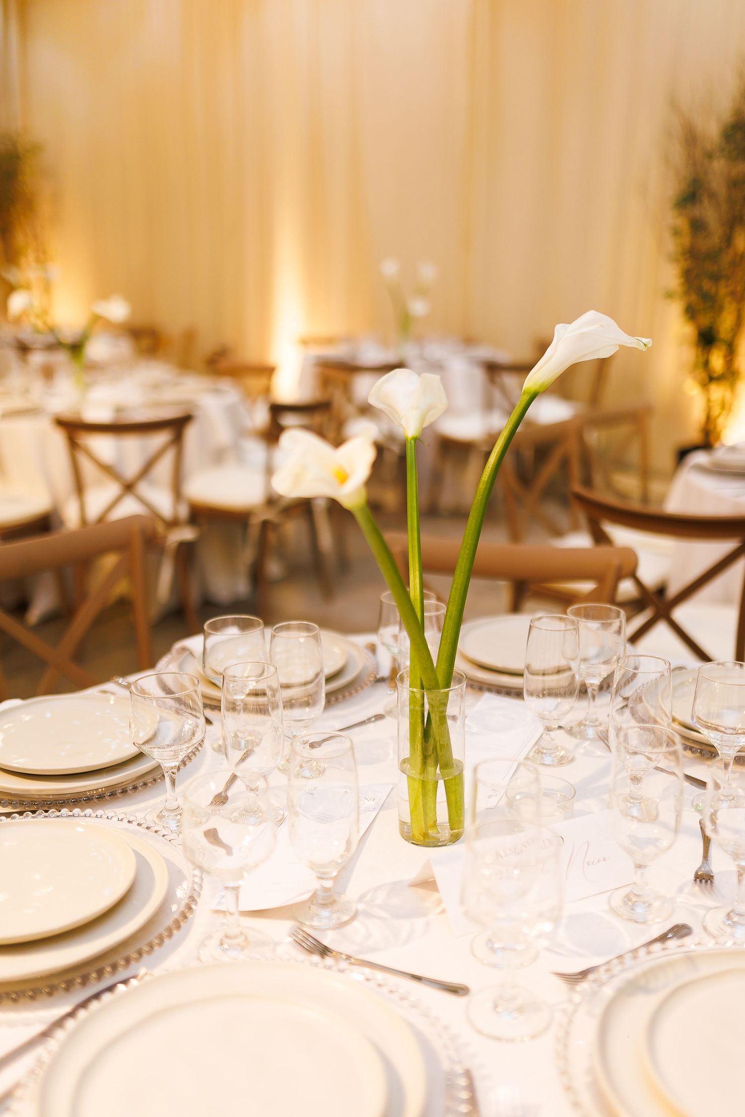 Elegant wedding reception table with white calla lilies centerpiece, crystal glasses, and white plates on a white tablecloth.