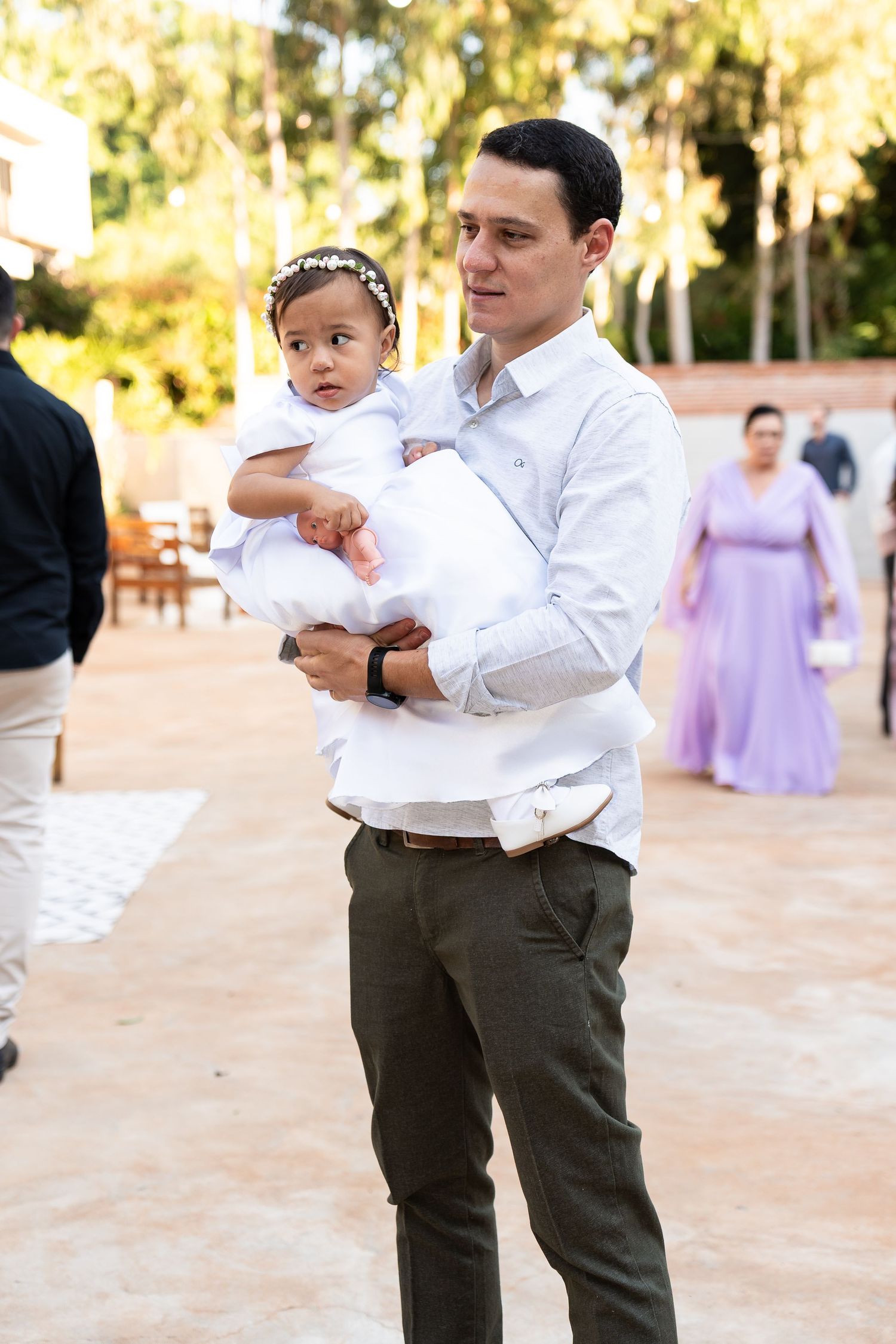 A man in a white shirt and olive pants holds a baby girl in a white dress at an outdoor event.