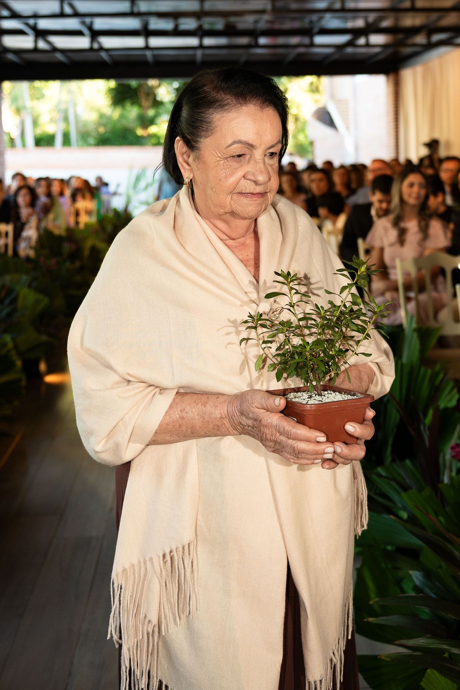 Elderly woman in cream shawl holding a potted plant at an indoor event with guests seated in background.