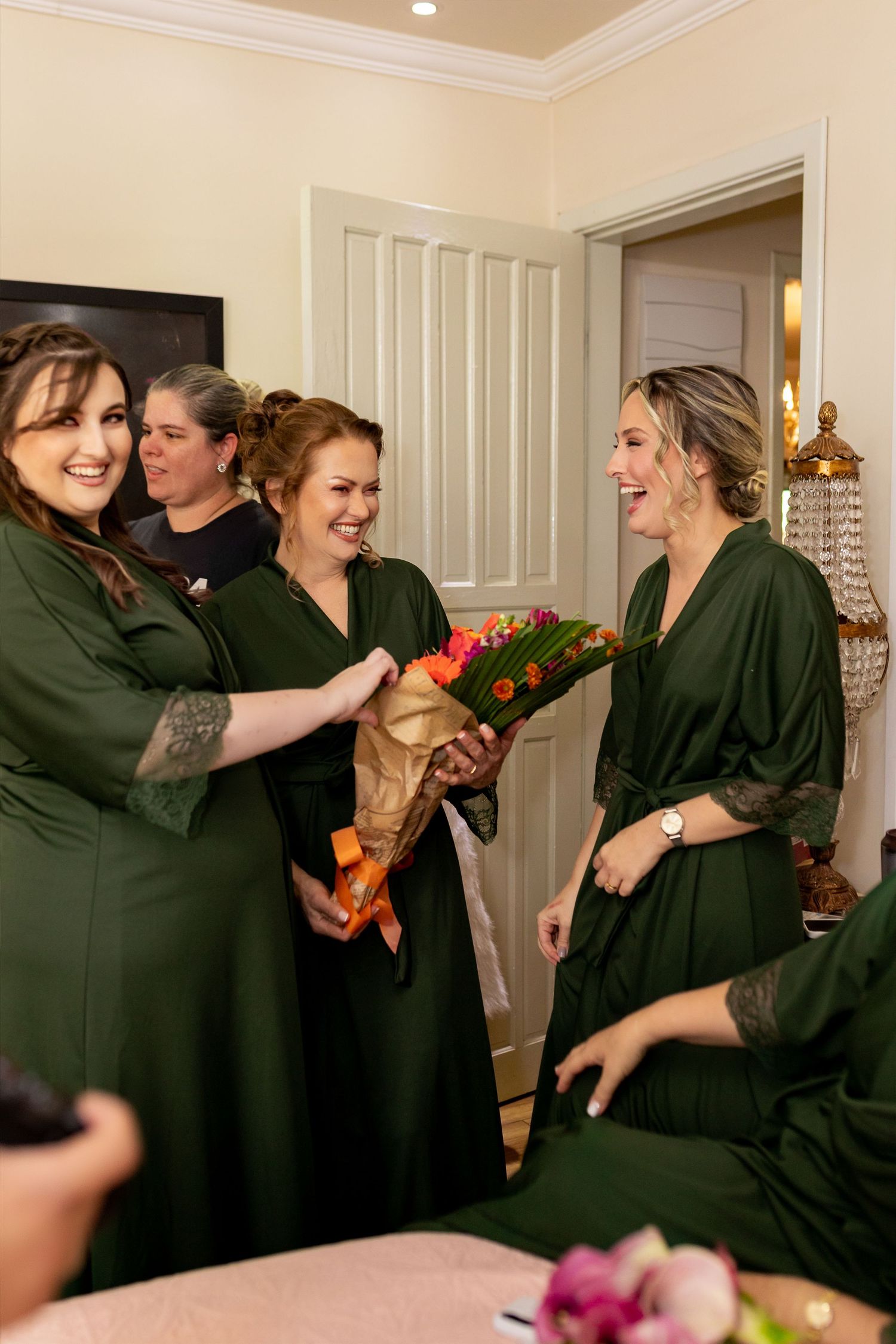 Bridesmaids in matching dark green robes laughing and sharing a colorful bouquet of flowers during wedding preparations.