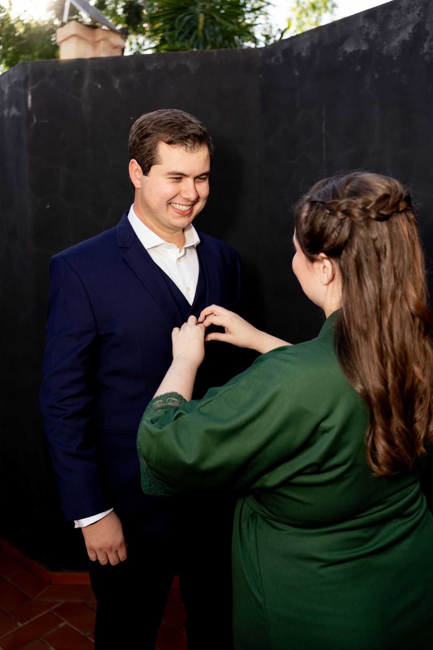 Woman in green dress adjusting boutonniere on smiling man in navy suit outdoors.