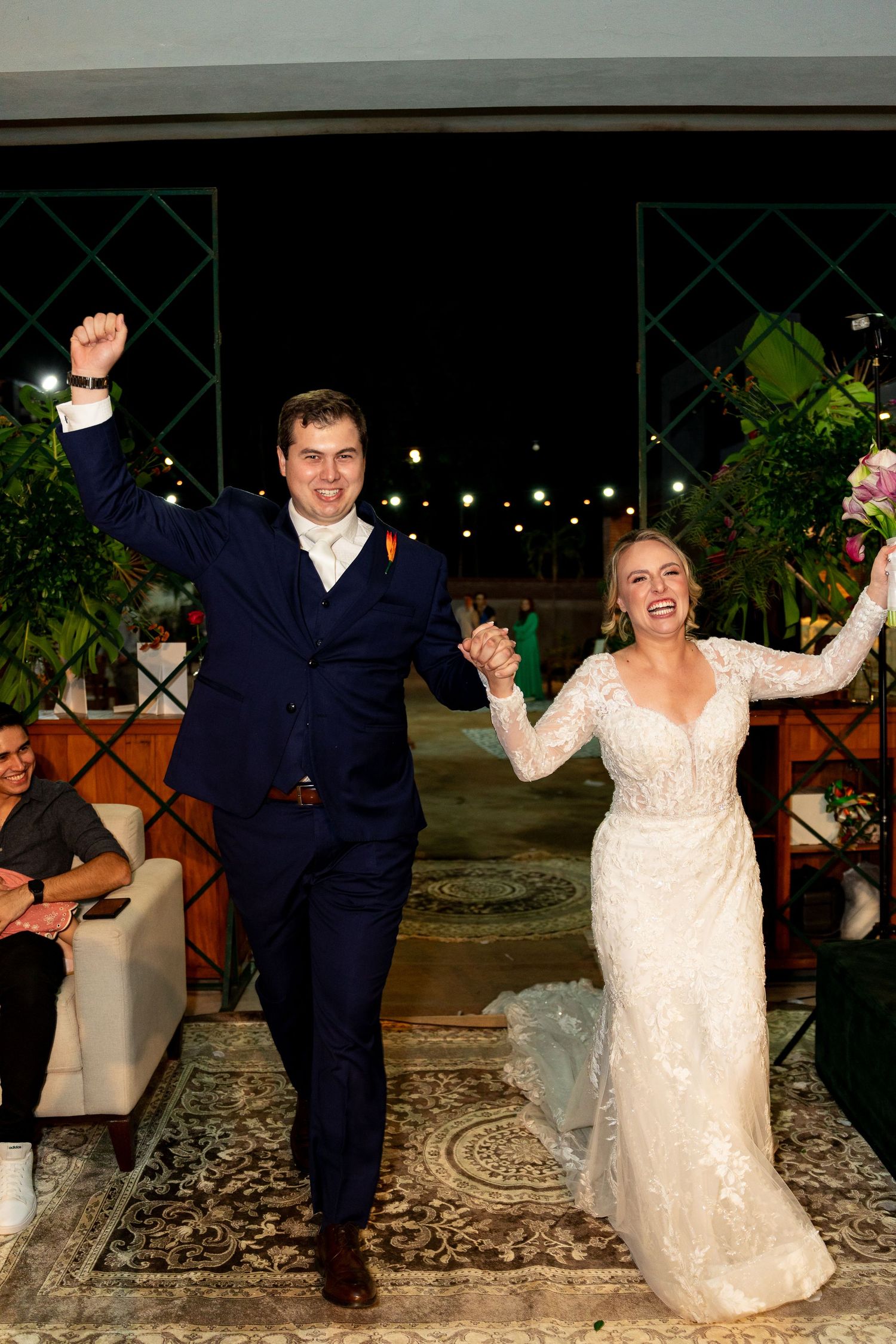 Joyful newlyweds celebrate their wedding reception entrance, bride in lace gown holding bouquet, groom in navy suit.