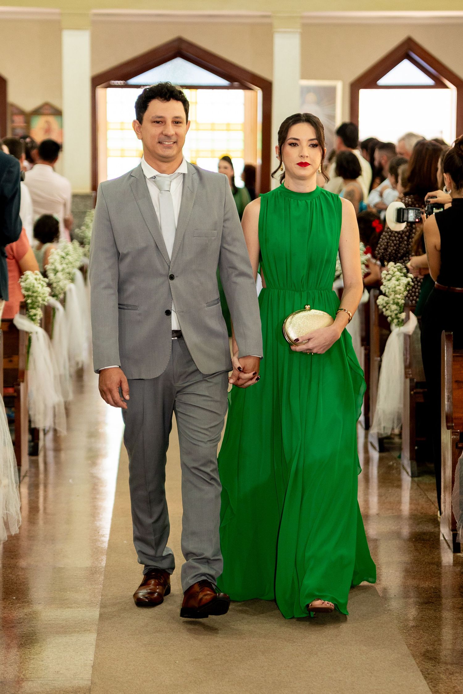 Couple walking down church aisle, man in gray suit, woman in elegant green gown, holding hands at wedding ceremony.