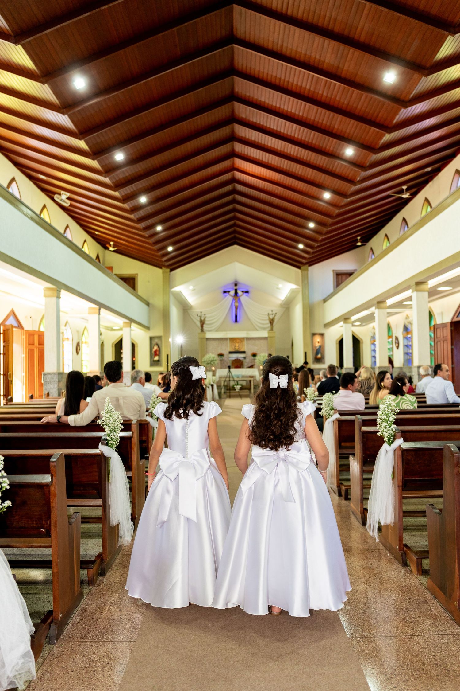 Two flower girls in white dresses walk down a church aisle during a wedding ceremony.
