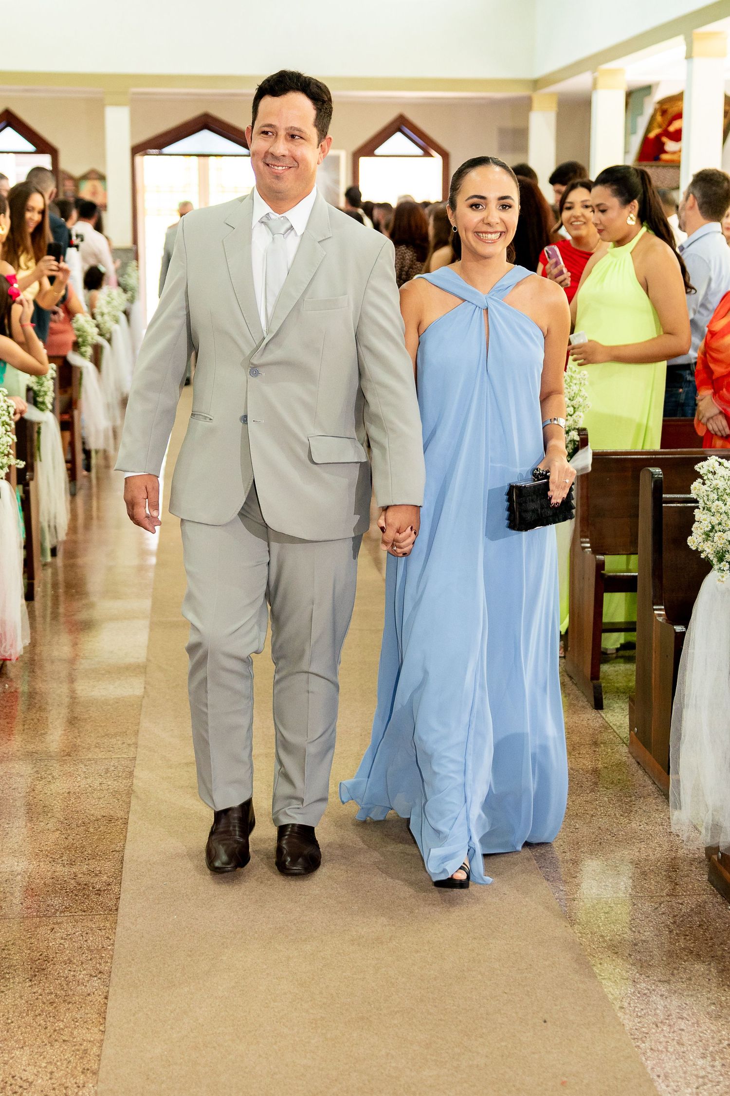 Couple walking down church aisle, man in gray suit, woman in light blue halter gown, holding hands at wedding.