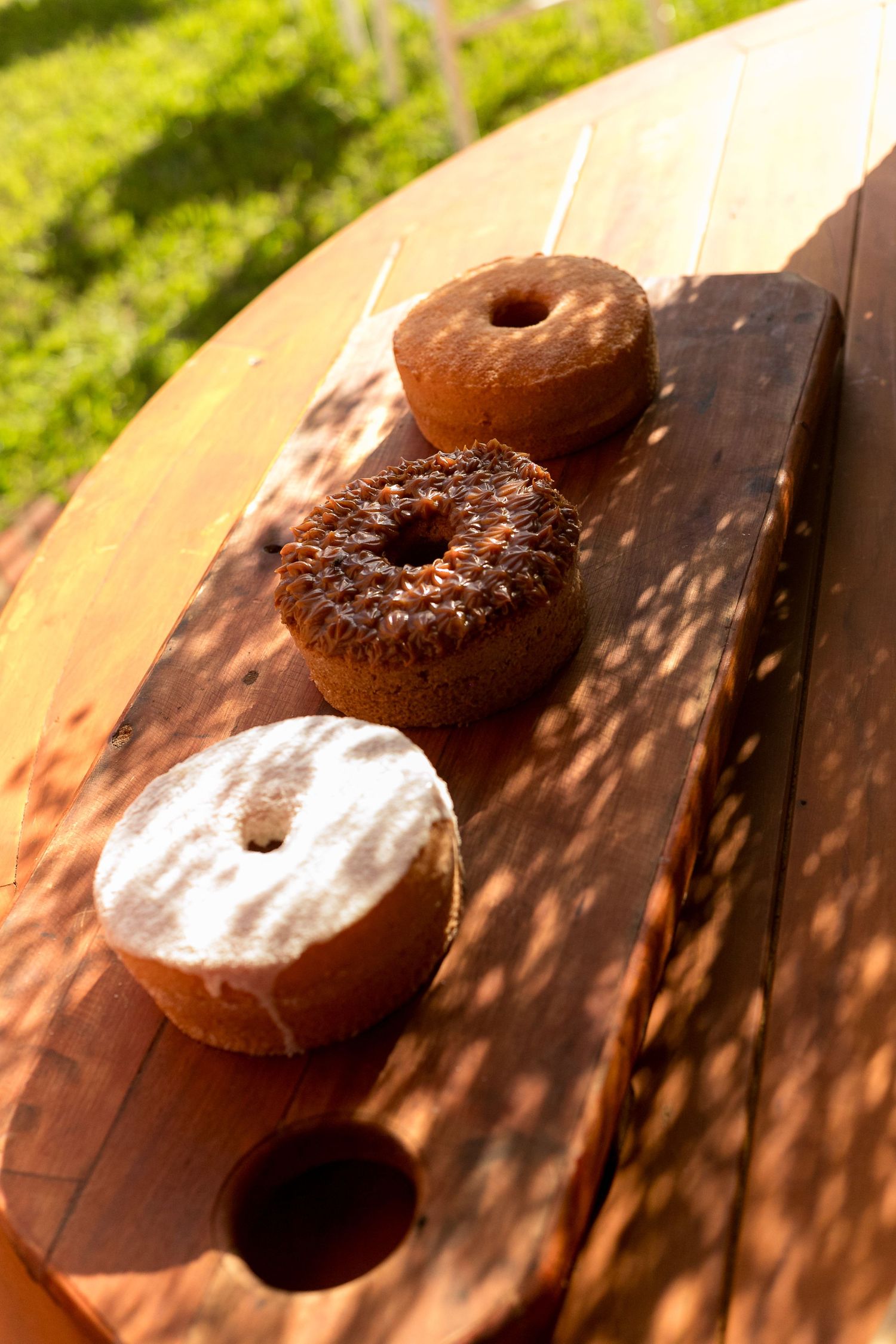 Three assorted donuts on a wooden cutting board outdoors in sunlight, including glazed, chocolate sprinkle, and powdered sugar varieties.