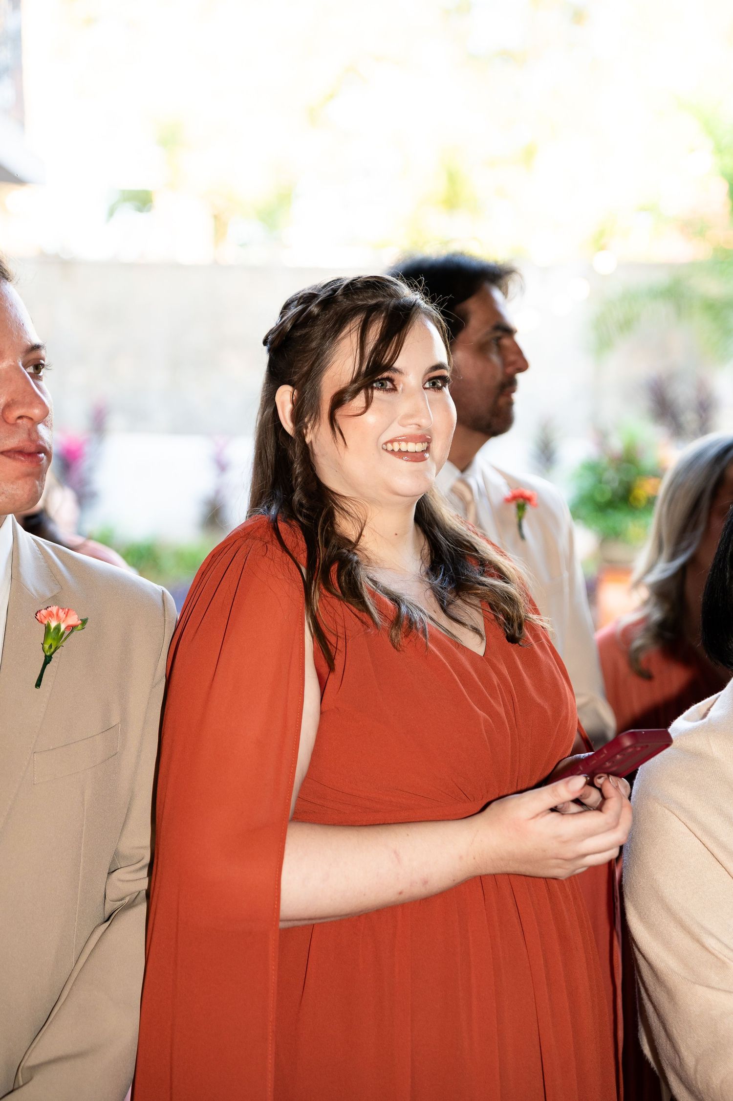 Woman in rust orange cape dress smiling at a wedding ceremony with guests in background.