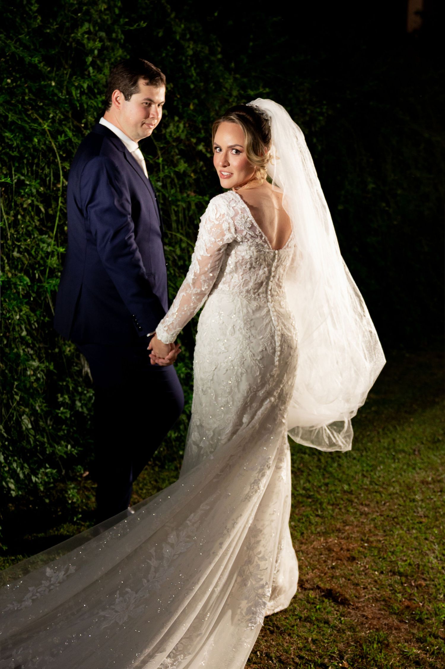 Bride in lace gown with veil and groom in navy suit hold hands outdoors at night near green hedges.