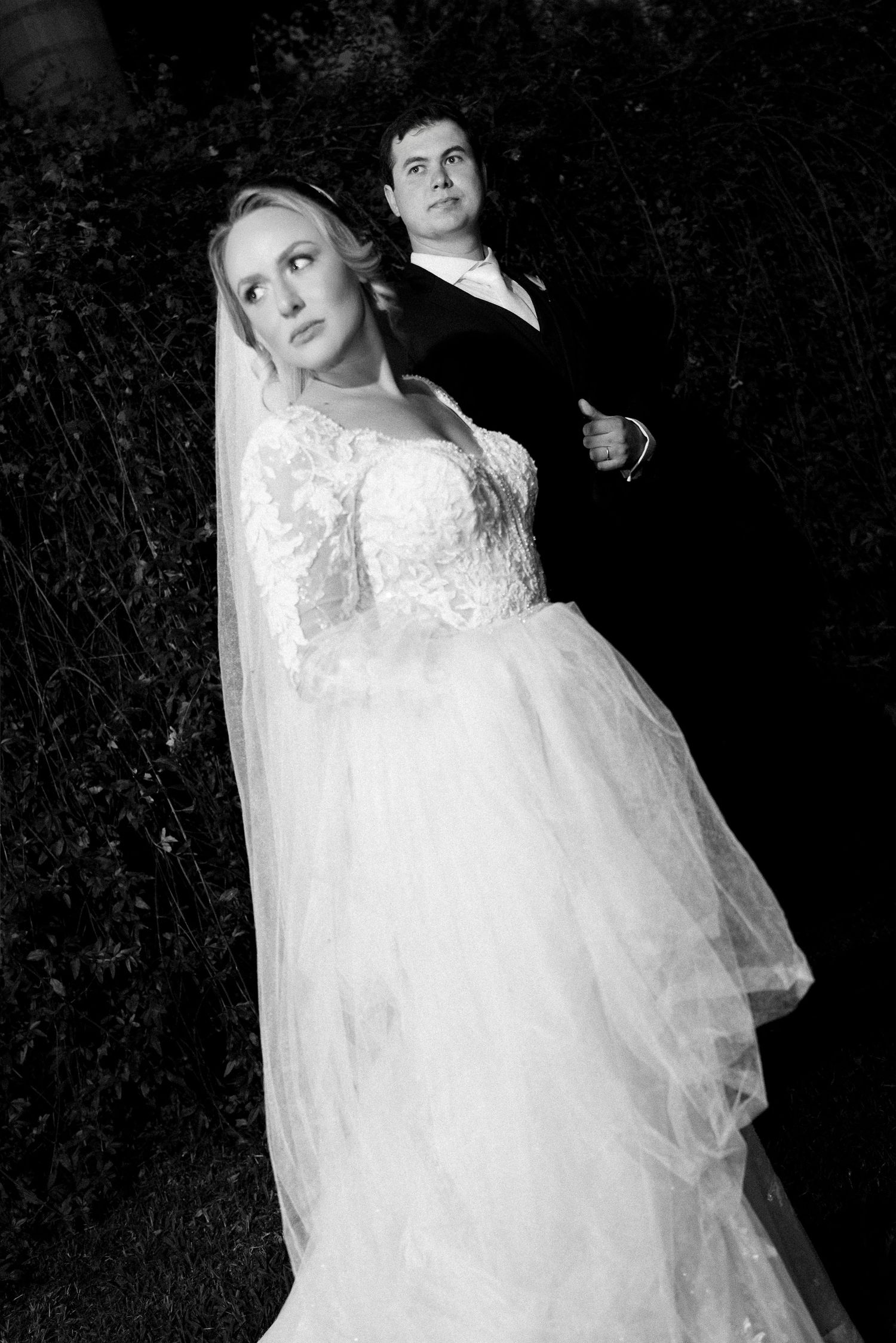 Black and white wedding photo of bride in lace gown with veil and groom in tuxedo posing outdoors at night.