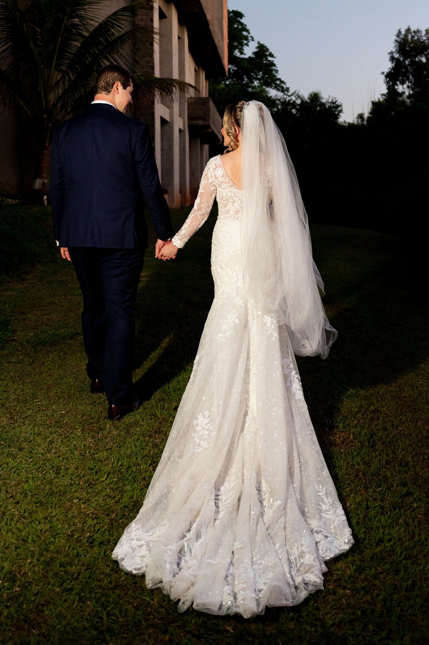 Bride in lace gown with veil holds hands with groom in navy suit walking outdoors at dusk.