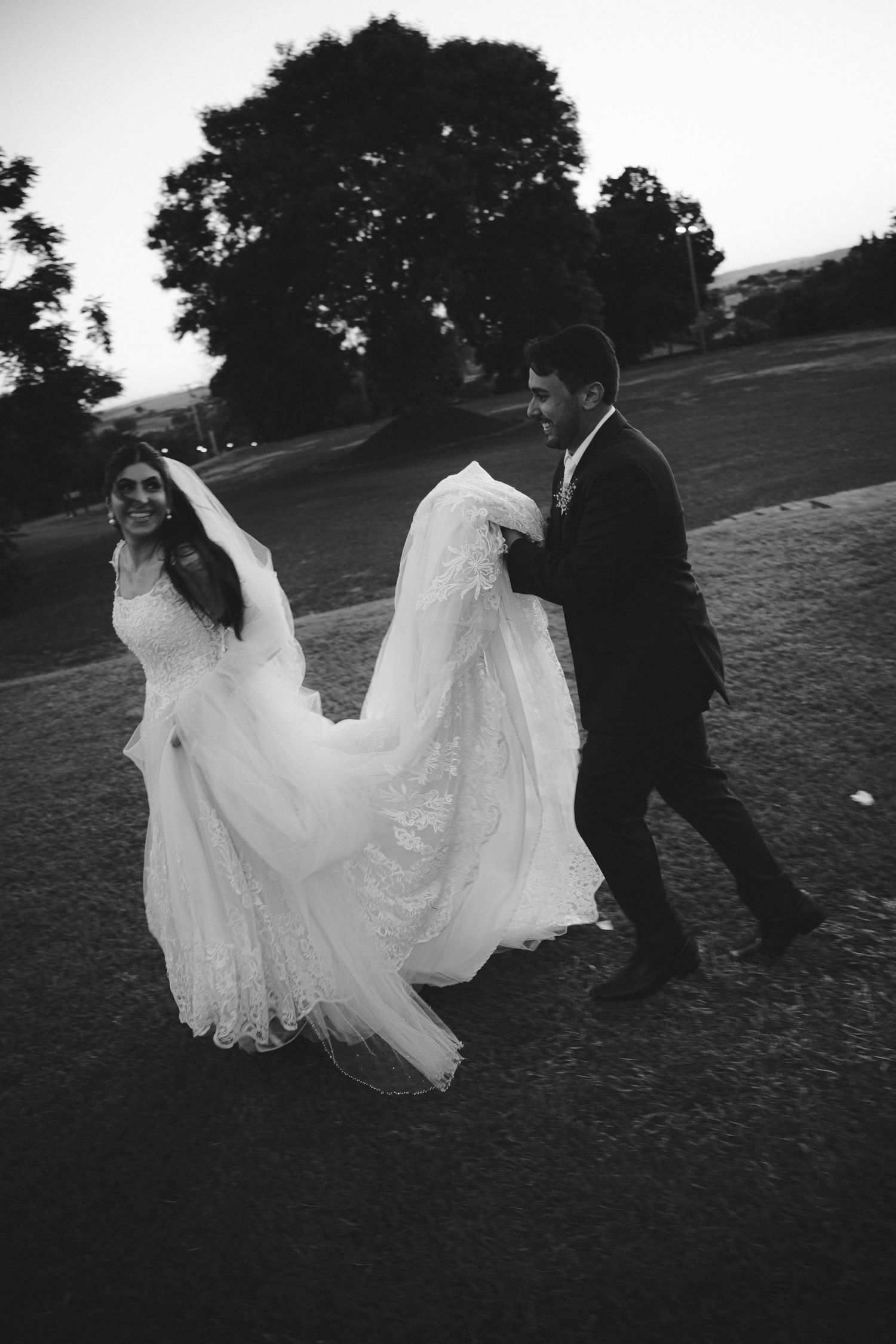 Black and white photo of a bride in a lace gown and groom in a suit walking outdoors at dusk.