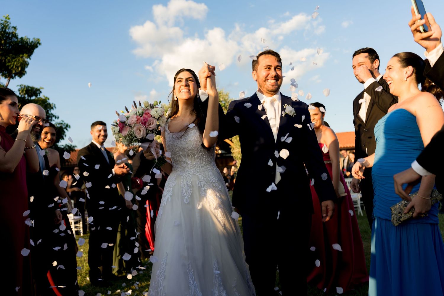 Bride and groom walk through confetti shower surrounded by guests at outdoor wedding ceremony.