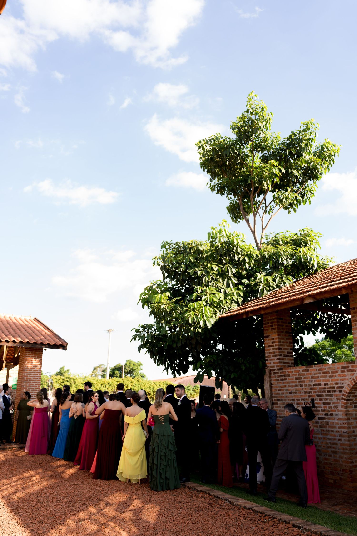 Guests in colorful formal attire gather outside a rustic brick venue surrounded by lush greenery under a blue sky.
