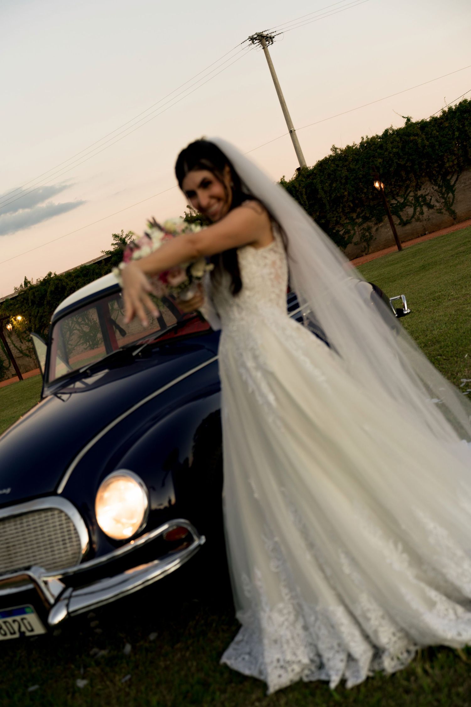 Bride in white lace wedding gown and veil poses with bouquet beside a classic dark blue vintage car at dusk.