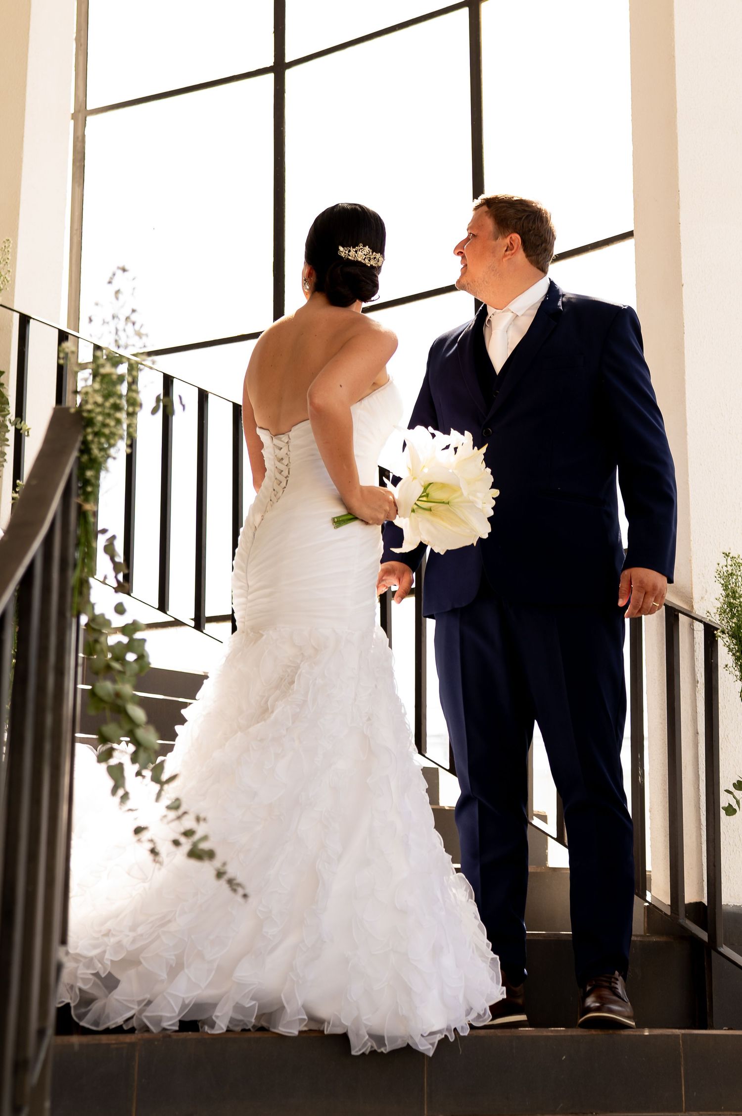 Bride in white mermaid gown and groom in navy suit share a romantic moment on stairs holding white lily bouquet.