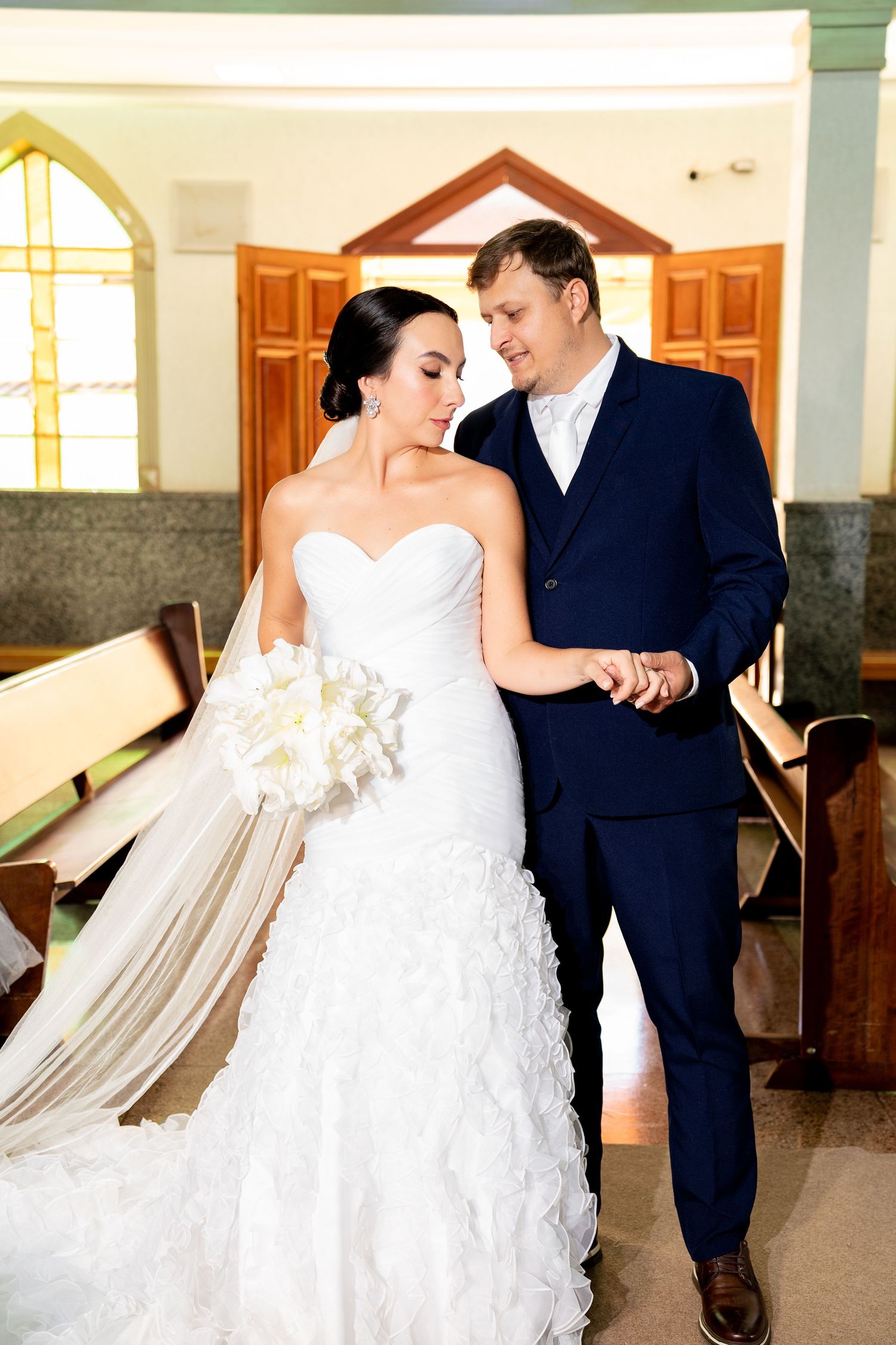 Bride in white strapless gown holding bouquet walks with groom in navy suit down church aisle on wedding day.