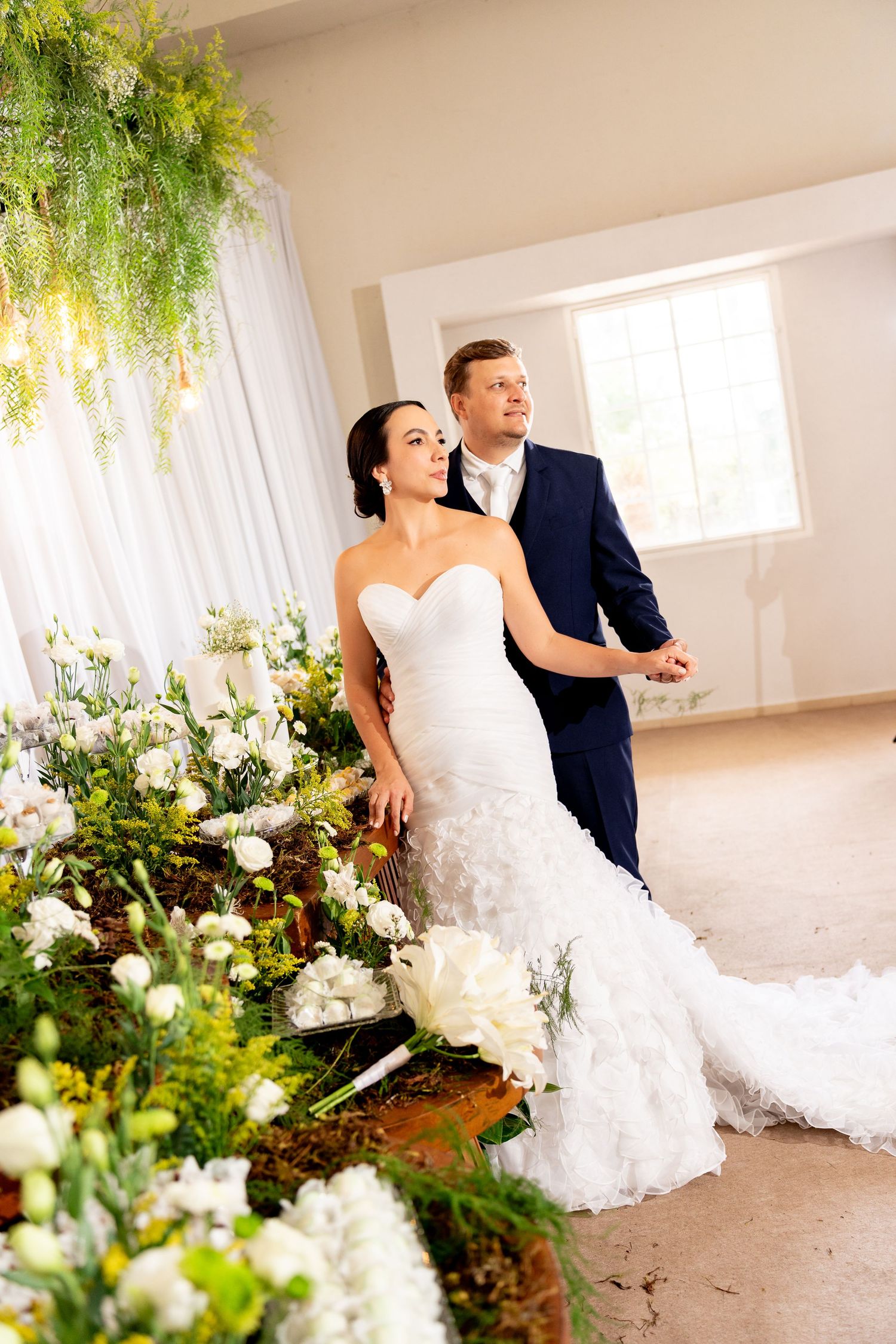 Bride in white mermaid gown and groom in navy suit pose elegantly among lush green floral arrangements indoors.