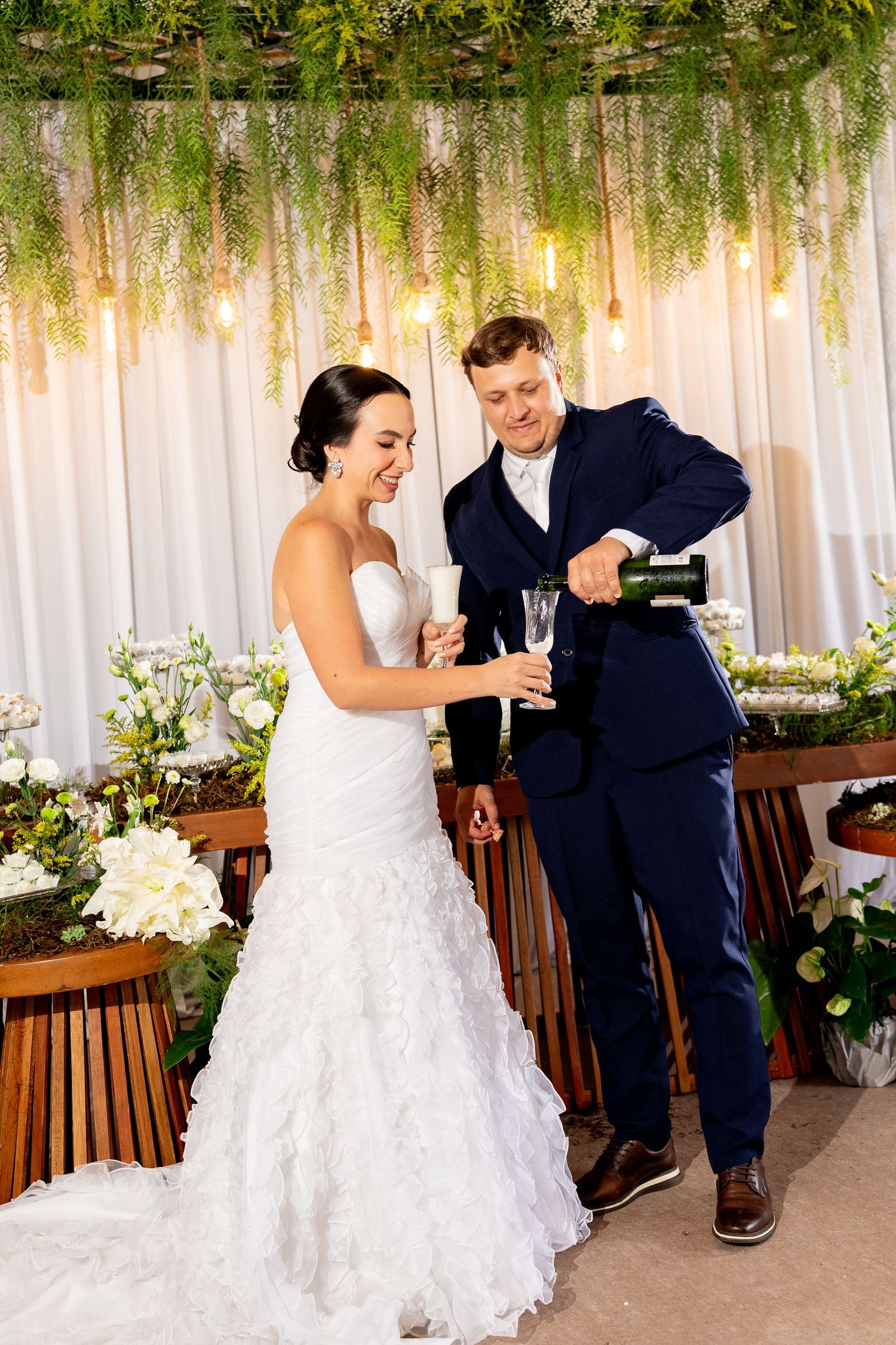 Bride and groom pouring champagne at wedding reception, decorated with greenery, flowers, and warm string lights.