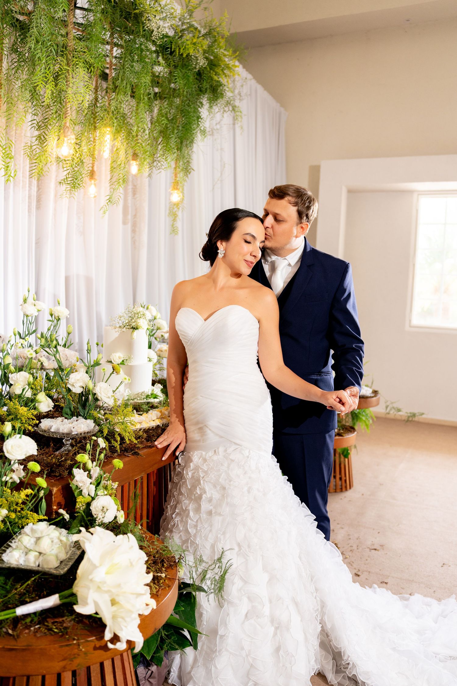 Bride in white mermaid gown and groom in navy suit embrace near floral arrangements at elegant wedding ceremony.
