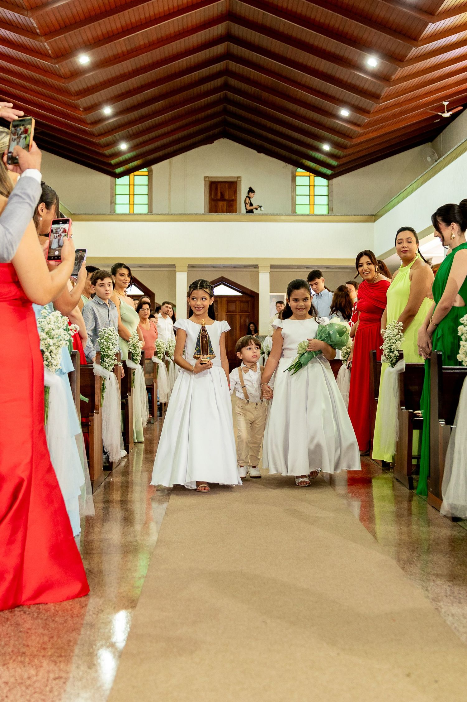 Two flower girls in white dresses walk down a church aisle during a wedding ceremony, flanked by guests in colorful attire.