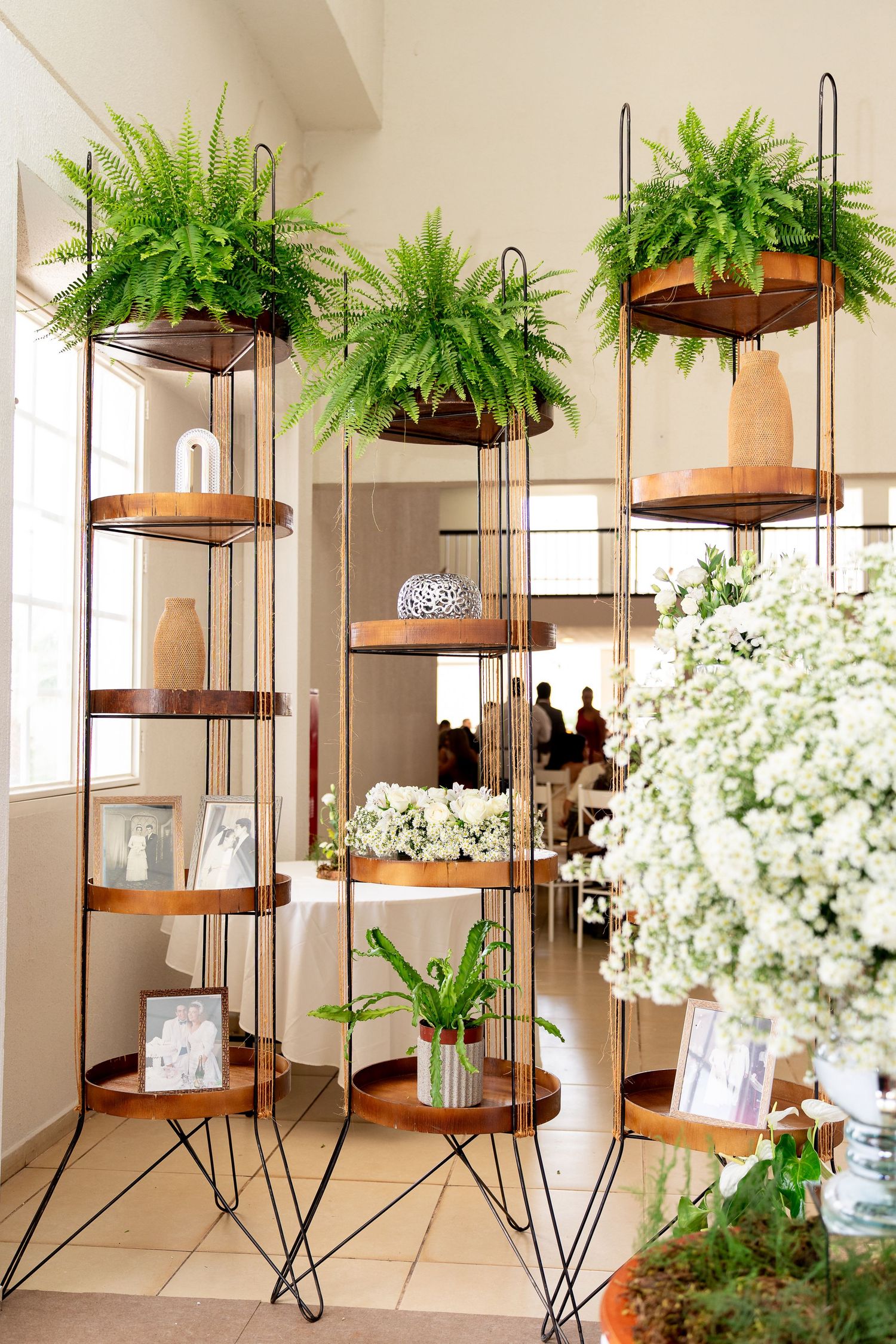Modern tiered metal shelving units with wooden shelves displaying ferns, white flowers, and decorative vases in a bright interior.