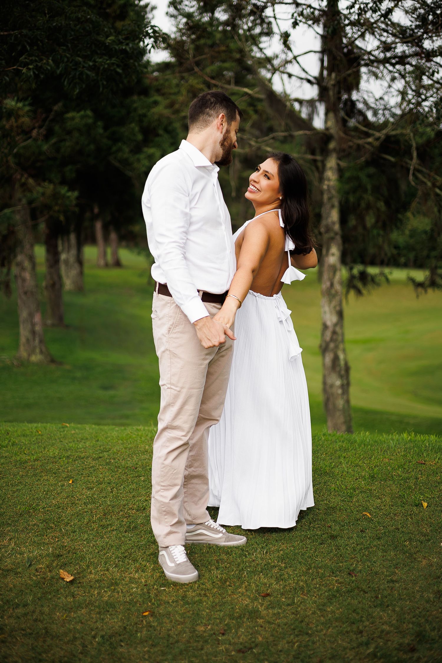 Romantic couple holding hands in a lush green park, woman in white backless dress smiling at man in white shirt.